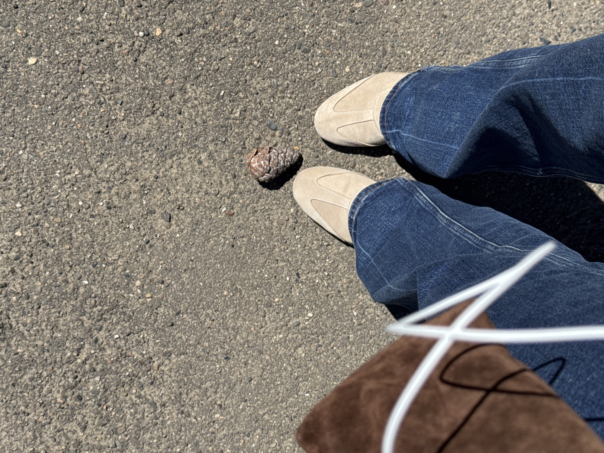 A high-angle shot on asphalt pavement shows a person's legs from the knees down, wearing blue jeans and tan-colored slip-on shoes. Between their feet is a single pine cone. A brown bag with white drawstrings is visible on the right side of the frame. The location cannot be determined from this image.