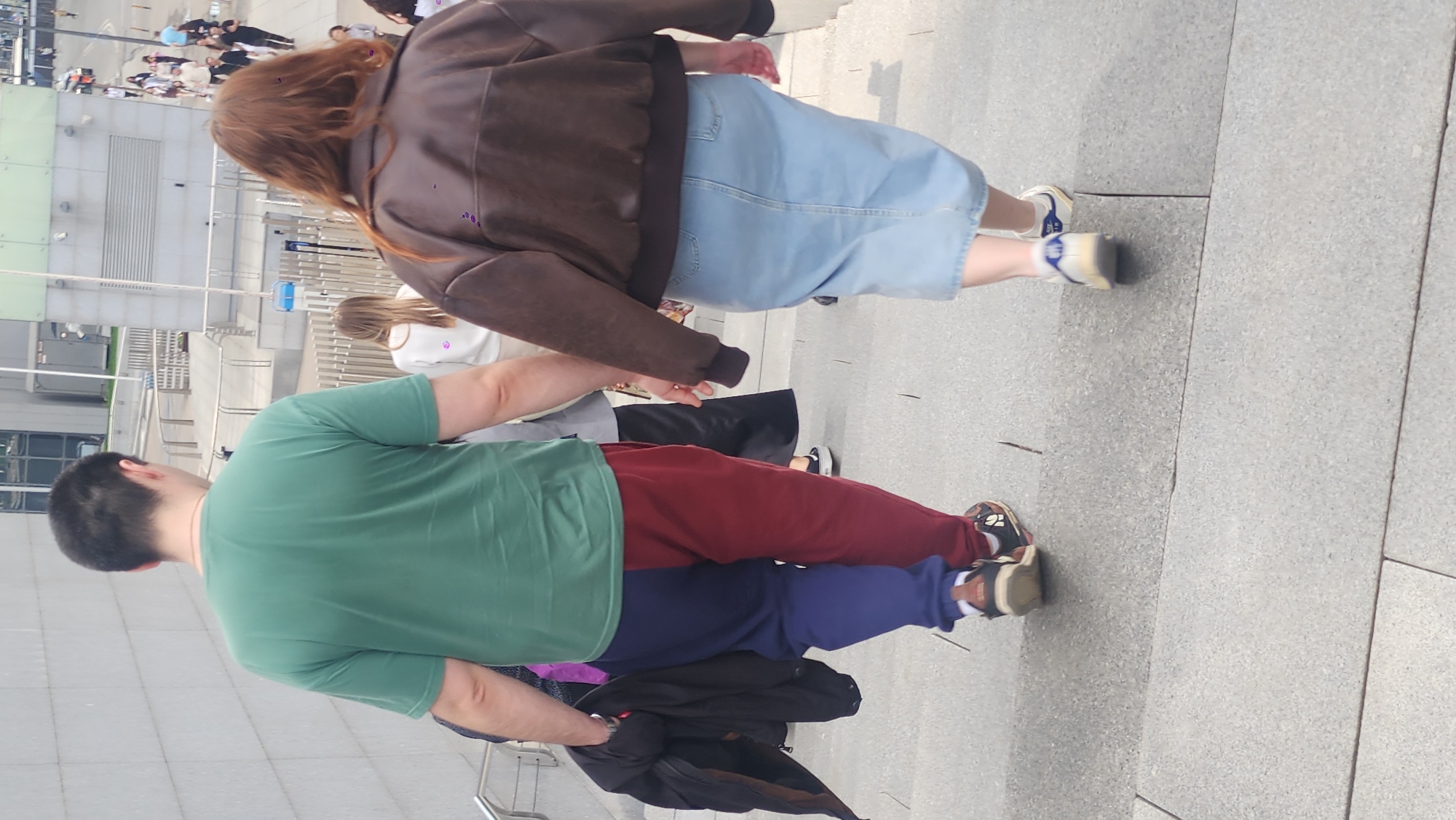 A group of people is seen walking, with two individuals in the foreground. One person wears a green t-shirt and two-toned pants, red and blue. Another person wears a brown jacket and a denim skirt. The location appears to be Moscow, Russia, based on architectural characteristics typical of the city.