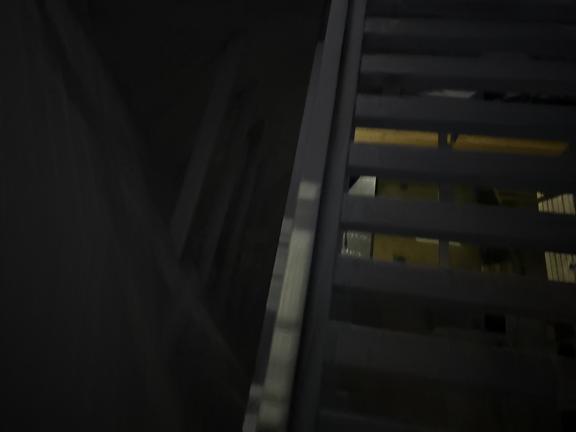The image depicts a dimly lit, indoor scene. In the foreground, a light-colored railing runs horizontally across the frame, with vertical slats visible beneath it. Beyond the railing, a yellow object is partially visible, suggesting some sort of appliance or structure. The background is dark and indistinct, with diagonal lines suggesting a sloped ceiling or roof. There are no people or discernible actions. The location is identified as Doma 1096 Km, Russia.
