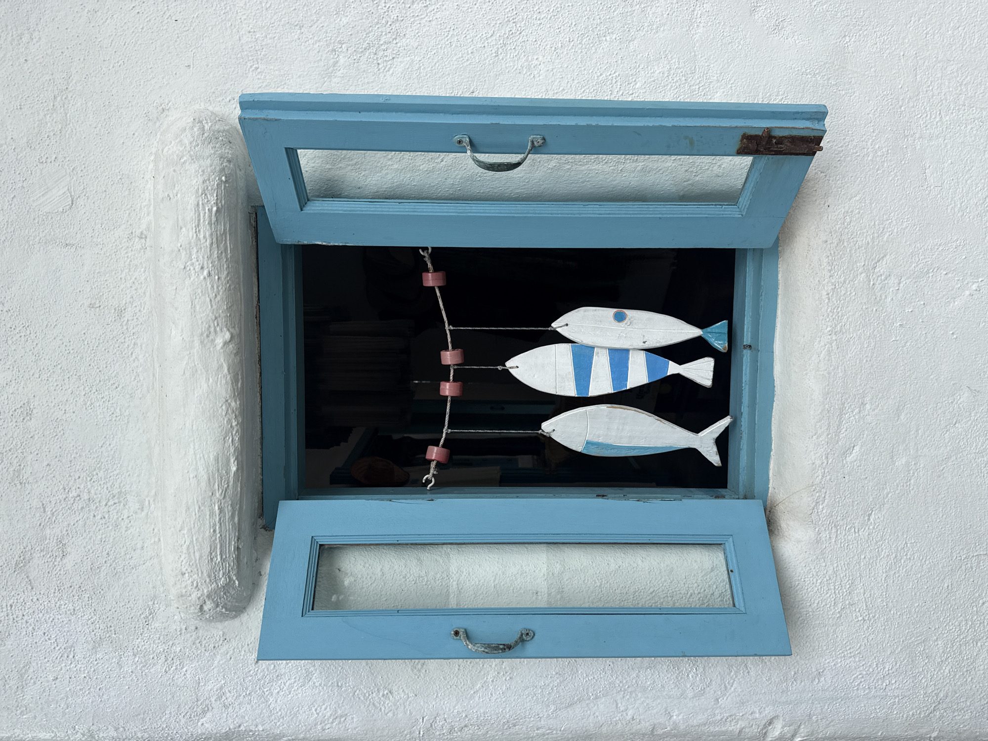 The image shows a close-up view of a white, textured wall with an open blue window. Inside the window, three wooden fish decorations are hanging from a string. The fish are white with blue accents. The left fish has a blue tail and a small blue circle on its body. The middle fish has blue stripes and a blue tail. The right fish is primarily white with blue accents and a blue tail. The window has visible metal handles and hinges, and the glass panes are partially obscured by a textured white material. The surrounding wall has a rough, stucco-like finish. The scene appears to be an exterior view of a building. The location is not specified within the image itself.