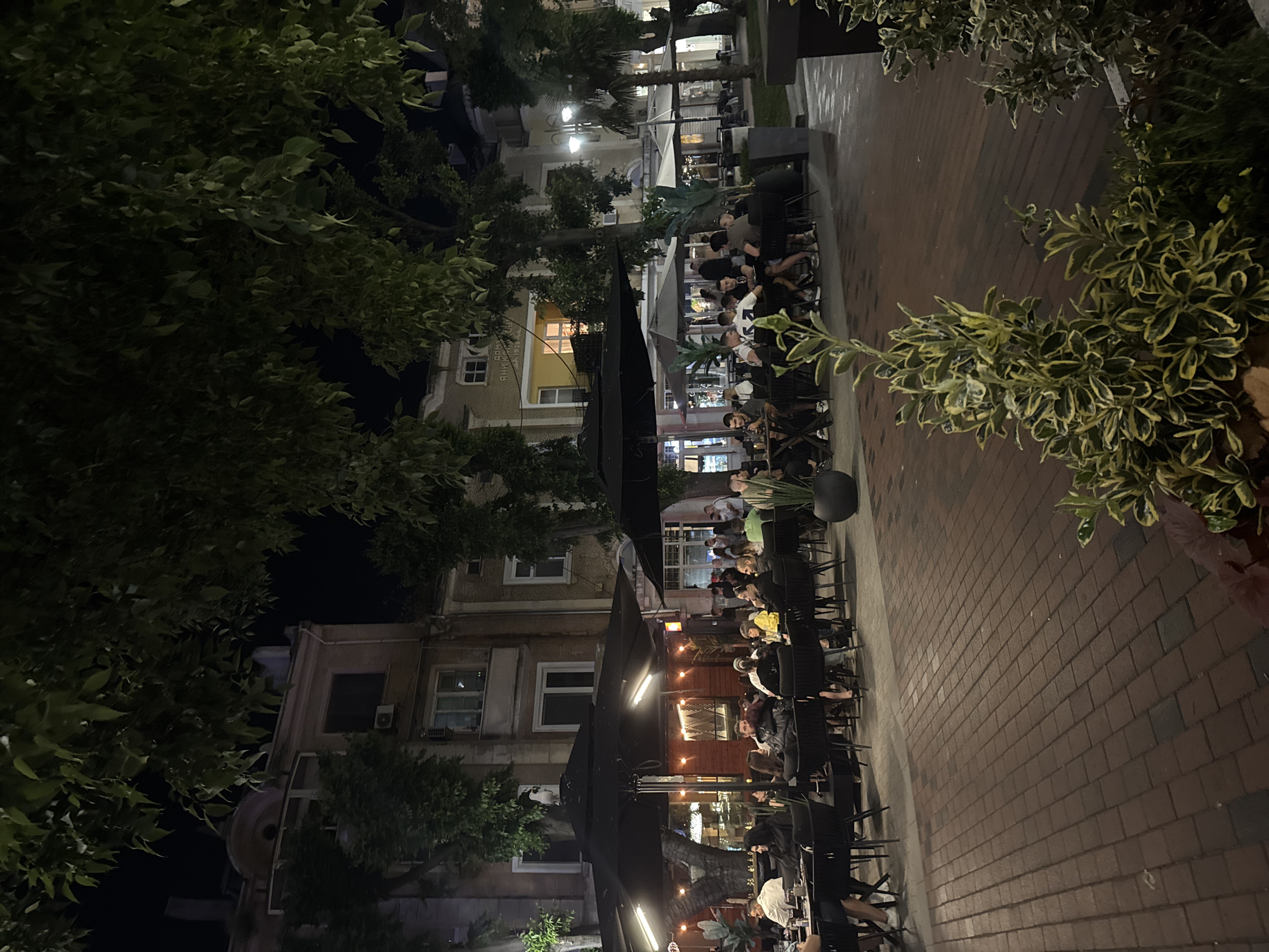 The image shows an outdoor seating area of a restaurant or cafe at night, located on a brick-paved pedestrian street in Varna, Bulgaria. Several black umbrellas provide shade over tables where groups of people are seated. The patrons appear to be engaged in conversation and dining, with some holding drinks. The building facades visible in the background are illuminated, revealing architectural details and windows. Trees with lush foliage overhang the scene, contributing to a lively, evening ambiance. The overall impression is of a popular urban gathering spot in the city of Varna.
