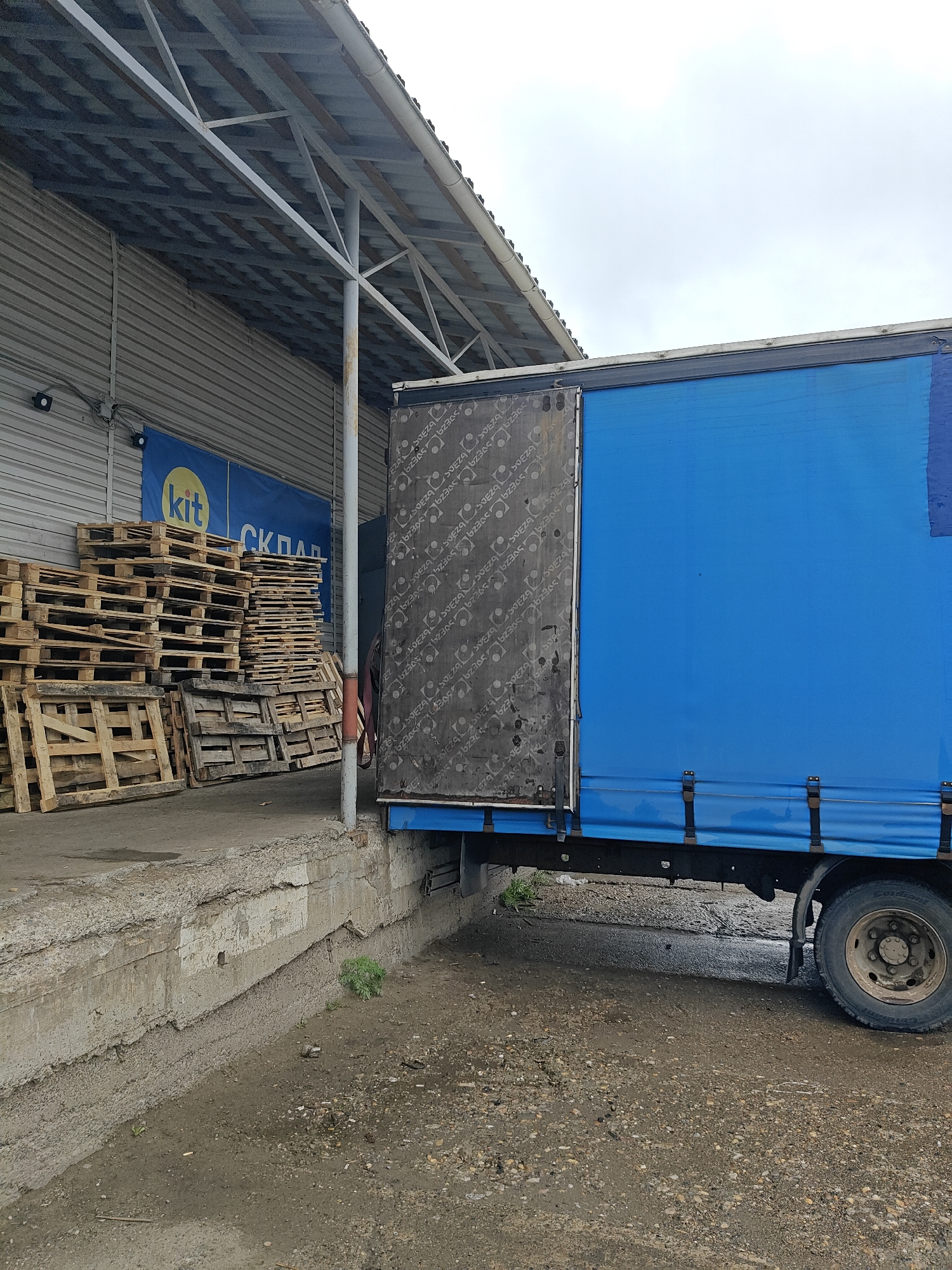 A medium shot shows a loading dock, a stack of pallets, and a blue semi-truck. The loading dock is made of concrete and has a worn, aged appearance. There are multiple stacks of wooden pallets to the left of the frame. A blue semi-truck is parked at the loading dock, with the back of its trailer facing the camera. The truck is a medium size, with a single set of wheels visible on the right side. The side of the trailer is made of a blue tarp material. The open rear door of the trailer is gray and appears to be made of wood. The environment is urban, possibly an industrial or commercial area, and has an overcast sky. The scene is located in the city of Chaikovsky, Russia.