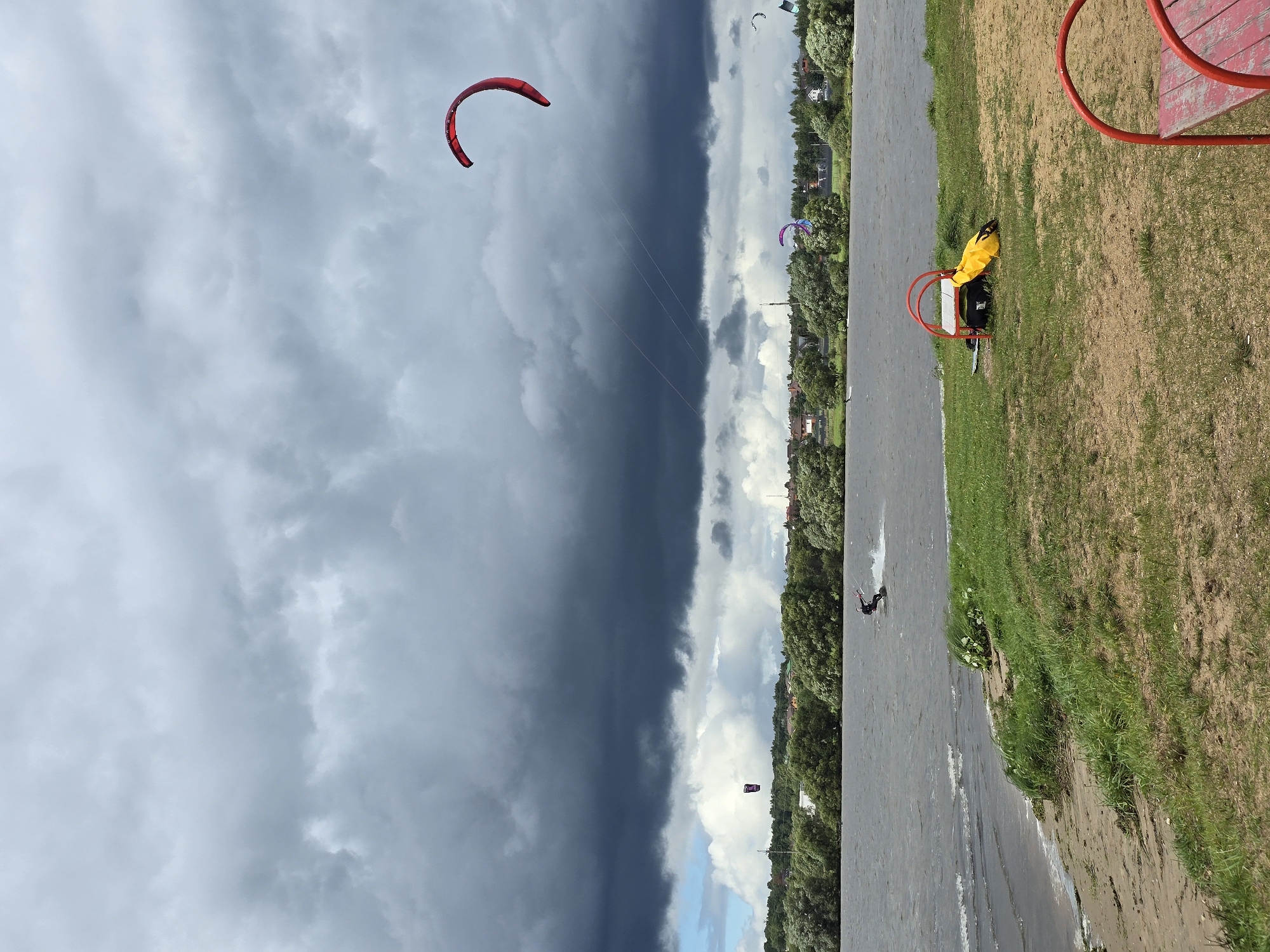 The image depicts a body of water, likely a lake or river, under a dramatic, cloudy sky. The water is choppy, indicating wind. On the water, a person is kitesurfing, actively engaged in the sport. In the background, a dense line of green trees and some residential buildings suggest a populated area. Several kites are visible in the sky, some appearing to be part of the kitesurfing activity, with one large red kite prominent in the upper right of the frame. The foreground shows a grassy embankment leading down to a sandy shore. On the grass, near the shore, is a small red playground structure with a yellow bag placed beneath it, implying a recreational area. The overall scene suggests an outdoor activity taking place in a scenic natural environment with signs of nearby civilization. The location is identified as Novoseltsevo, Russia.