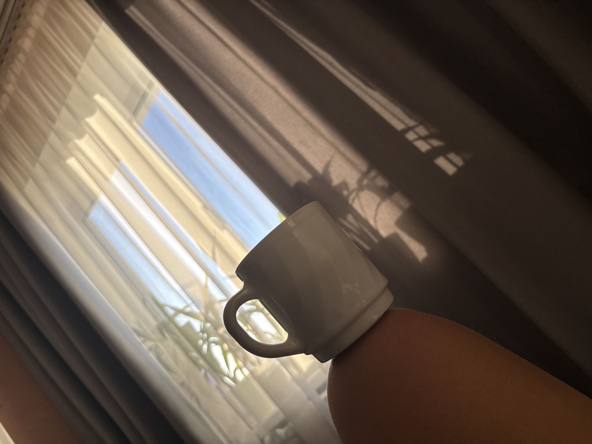 A white ceramic mug sits balanced on a person's knee. Behind the mug, light filters through two layers of curtains: sheer white curtains closest to the window and heavier brown curtains further back. The window reveals a view of blue sky. The photo was taken in a residential interior space in the city of Chaikovsky, Perm Krai, Russia.