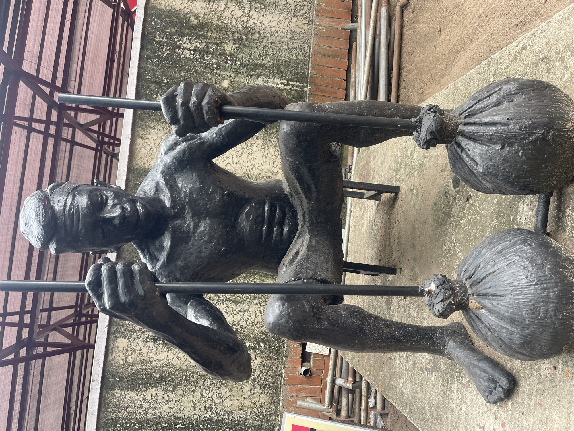 A ground-level, outdoor shot depicts a statue of a person suspended by their hands and feet from two metal bars. The person has a gaunt appearance. Large, sack-like objects are attached to the bars at the feet. The background is a concrete surface with metal pipes and scaffolding. The location is Unknown, Unknown.