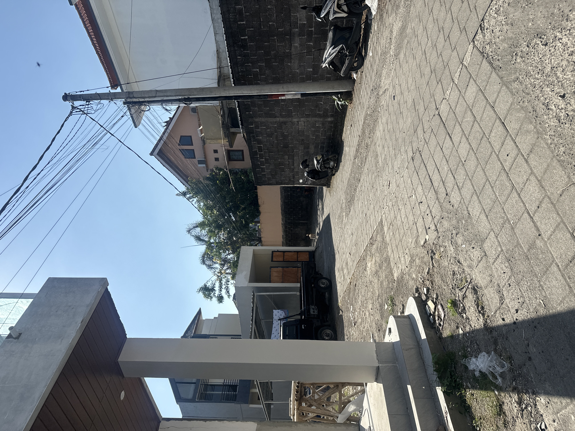 A black pickup truck is parked on a cobblestone street in Denpasar, Indonesia. A light-colored dog is walking on the street in the distance. The street is lined with a concrete wall and several houses. The sky is clear and blue.