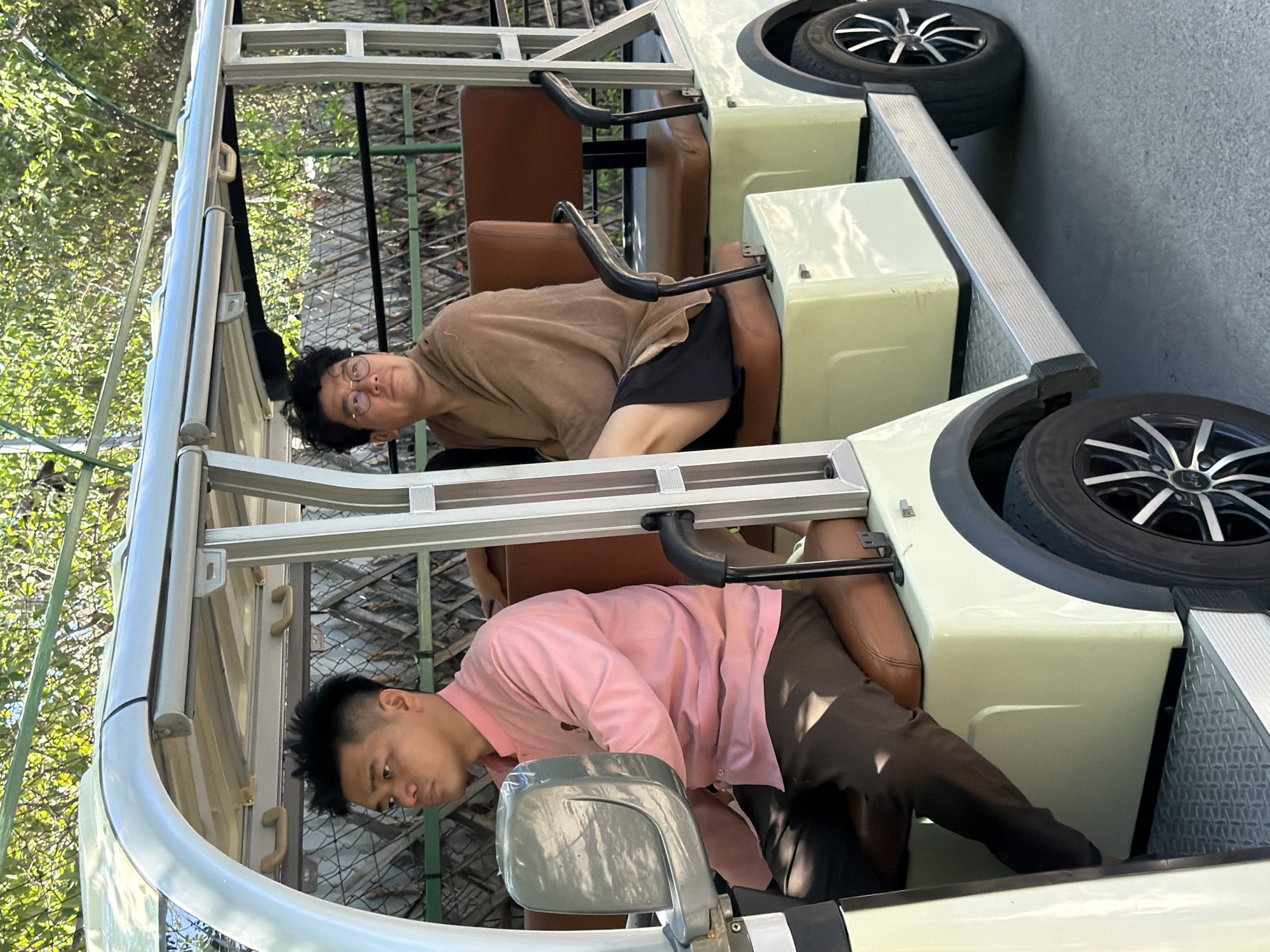 Two men are seated in a light-colored electric golf cart. The man on the left, wearing a pink collared shirt and dark pants, is driving. The man on the right, wearing glasses, a light brown wrap, and black shorts, is looking to his left. The golf cart has brown leather seats and is parked outdoors, with lush green trees and foliage in the background, suggesting a tropical environment. The ground appears to be a paved surface, likely a road or pathway. The overall impression is a casual outing in a sunny location.