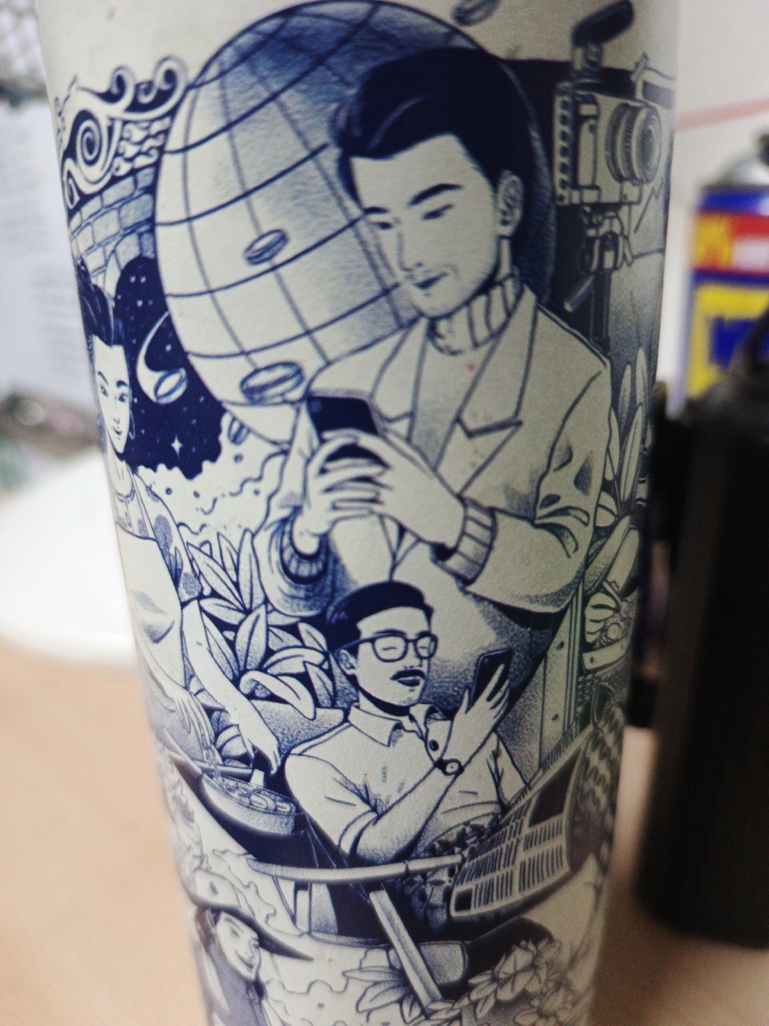 A blue and white illustration on a cylindrical object depicts several figures engaged with smartphones. In the background, a large globe is visible, with a retro camera on a tripod to the right. A man in the upper portion of the illustration is holding a smartphone and looking down at it. Below him, another man wearing glasses and a mustache is also looking at his phone. Further down, a woman with her hair pulled back is shown. The artwork is intricate and detailed, with elements of swirling patterns and foliage integrated into the composition. The background is blurred, suggesting the object is placed on a surface in an indoor setting, possibly a table. The object is likely a cup or a container given its shape and the nature of the artwork.