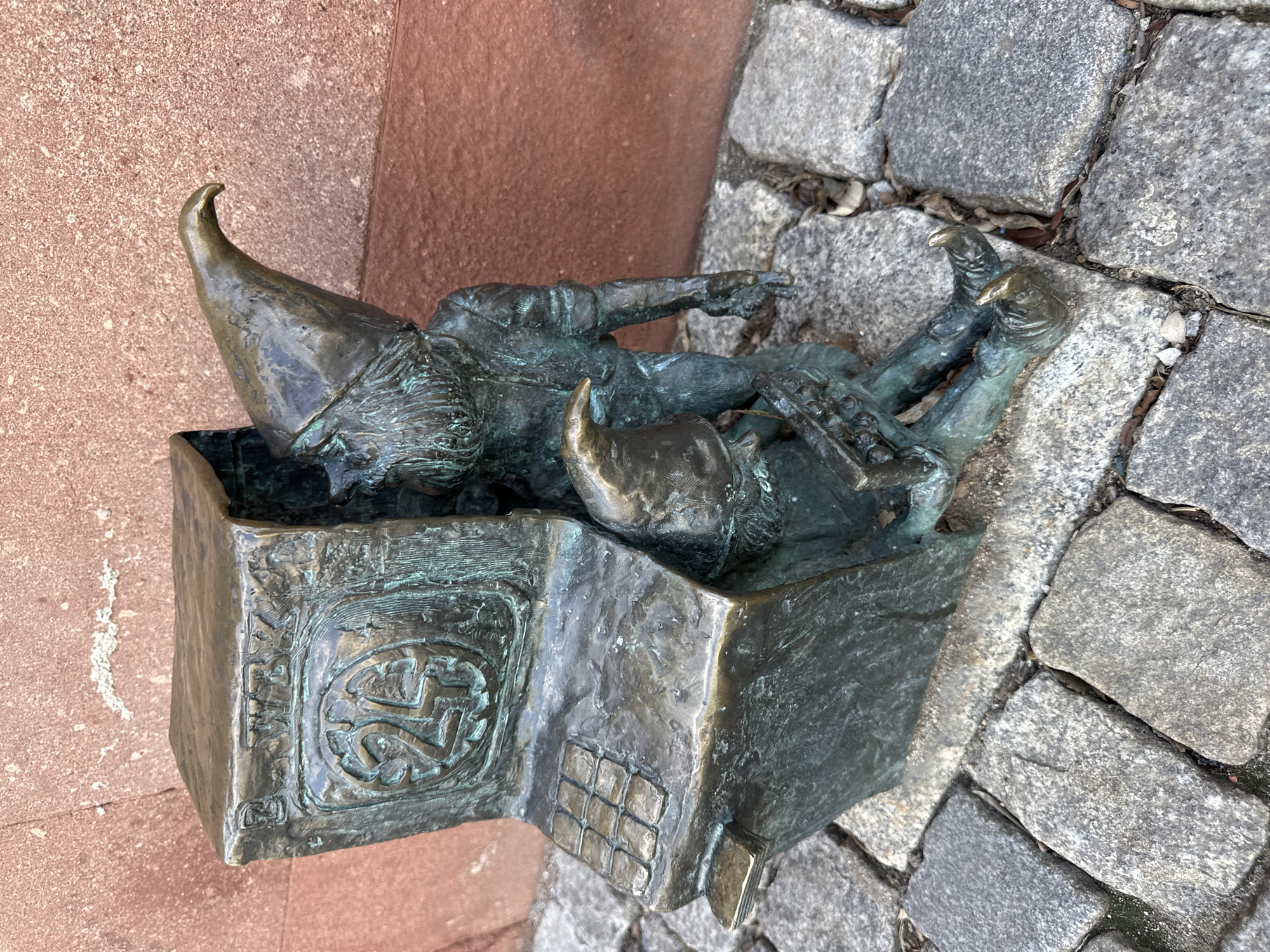 The image shows a bronze sculpture of two gnomes in a box, situated on a cobbled street next to a red brick wall. One gnome lies in the box, pointing, while the other is partially in the box, with his head and pointed hat visible. The box has detailed patterns on its side, including a symbol and grid-like structure. The statue is located in Wrocław, Poland, known for its gnome statues.