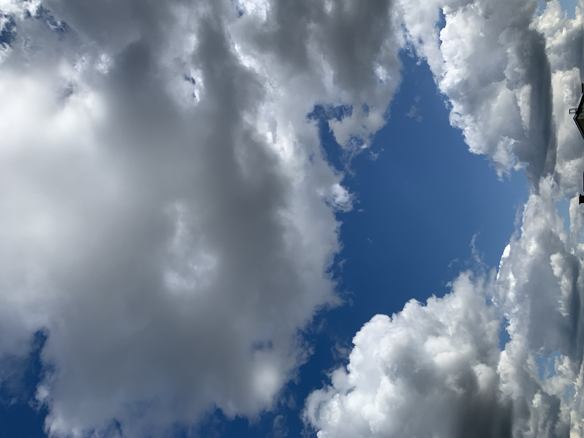 The image shows a blue sky with dramatic, varied clouds. Some clouds are bright white and fluffy, while others are darker gray and more ominous, suggesting potential for rain or storm. The clouds fill most of the frame, with patches of clear blue sky visible between them. Towards the bottom of the image, a sliver of rooftops and structures, possibly part of a residential area, can be seen against the horizon. The scene appears to be an outdoor view, likely taken from ground level looking upwards. No people or specific landmarks of Kapyĺ, Belarus are identifiable in this generic sky view.