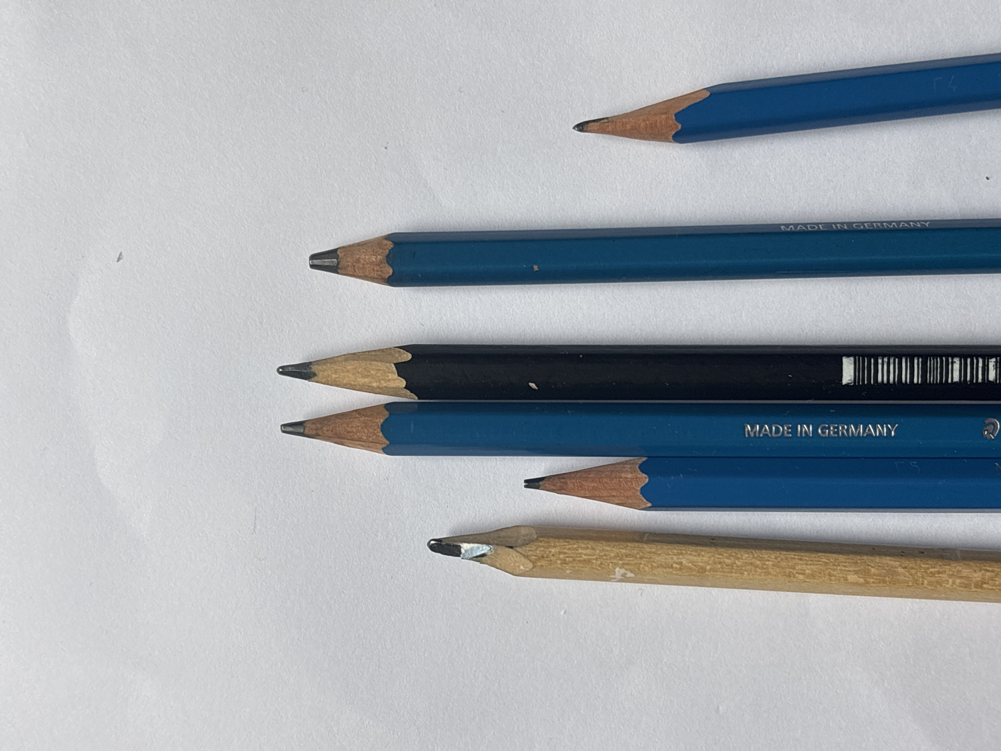 The image shows six pencils arranged on a white surface. The pencils are oriented horizontally, with the points facing left. From top to bottom, the pencils are: 1. A blue pencil with the point slightly out of focus. 2. A blue pencil with the inscription "MADE IN GERMANY". 3. A black pencil with a barcode. 4. A blue pencil with the inscription "MADE IN GERMANY". 5. A blue pencil. 6. A light brown, unpainted wooden pencil. The surface appears to be a sheet of paper. The scene is devoid of human subjects, actions, or environmental context suggesting a location in Choeng Thale, Thailand.