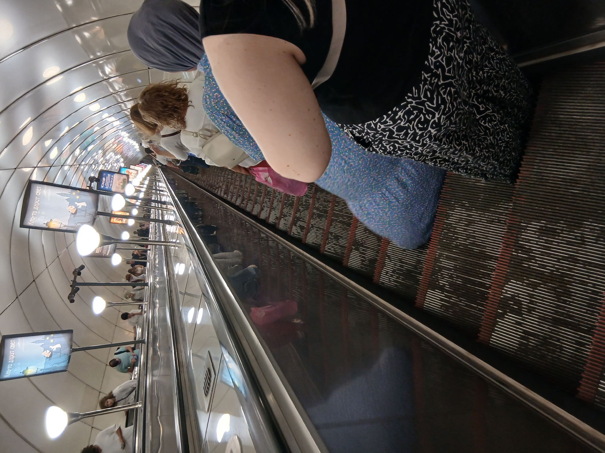 The image shows a group of people on an escalator in what appears to be a subway station. The escalator is moving upwards and is lined with reflective surfaces and lights. Several individuals are visible, some are standing and others are walking. One person in the foreground is wearing a black shirt and a black and white patterned skirt, and another person is wearing a blue patterned dress. There are posters or advertisements visible on the wall of the tunnel, with some text in what appears to be Cyrillic script. The lighting suggests it is daytime, but there are no windows to indicate the weather. The overall atmosphere seems functional and utilitarian, typical of public transportation infrastructure.