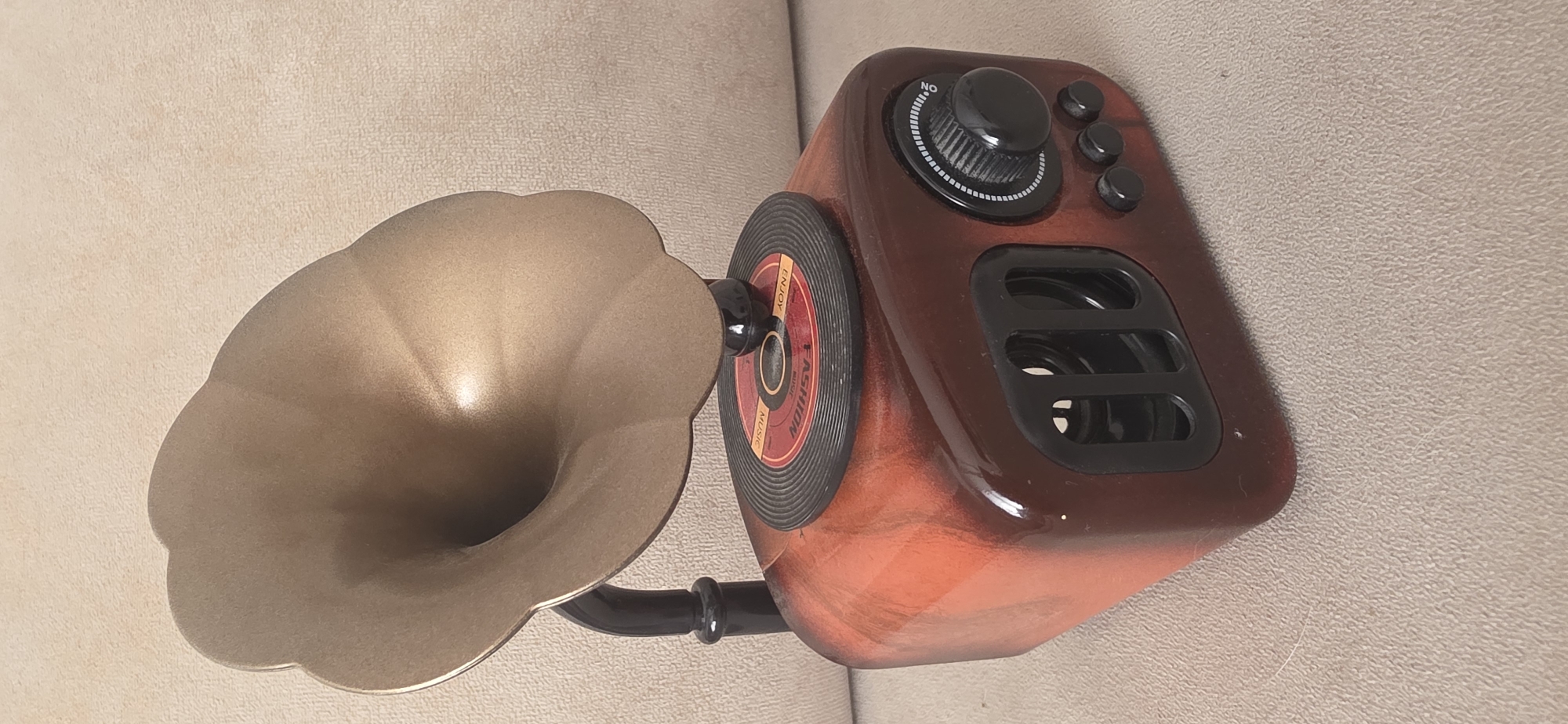 The image shows a close-up, eye-level, static shot of a decorative retro-style gramophone speaker on a textured beige surface. The speaker is designed to resemble an antique gramophone with a wooden-tone base, a gold-colored horn, and a miniature vinyl record on top. The base has a faux wood grain finish in shades of orange and brown. On the front of the base, there are two rectangular vents. On the right side of the base, there is a black rotary dial with white markings and a black knob, as well as three small black buttons. The gramophone horn is flared and made of a metallic gold-colored material. It is supported by a black arm attached to the base. The miniature vinyl record is black with a red label in the center. The overall impression is of a decorative electronic device. There are no people or any indications of the city of Sumgayit, Azerbaijan in this image.