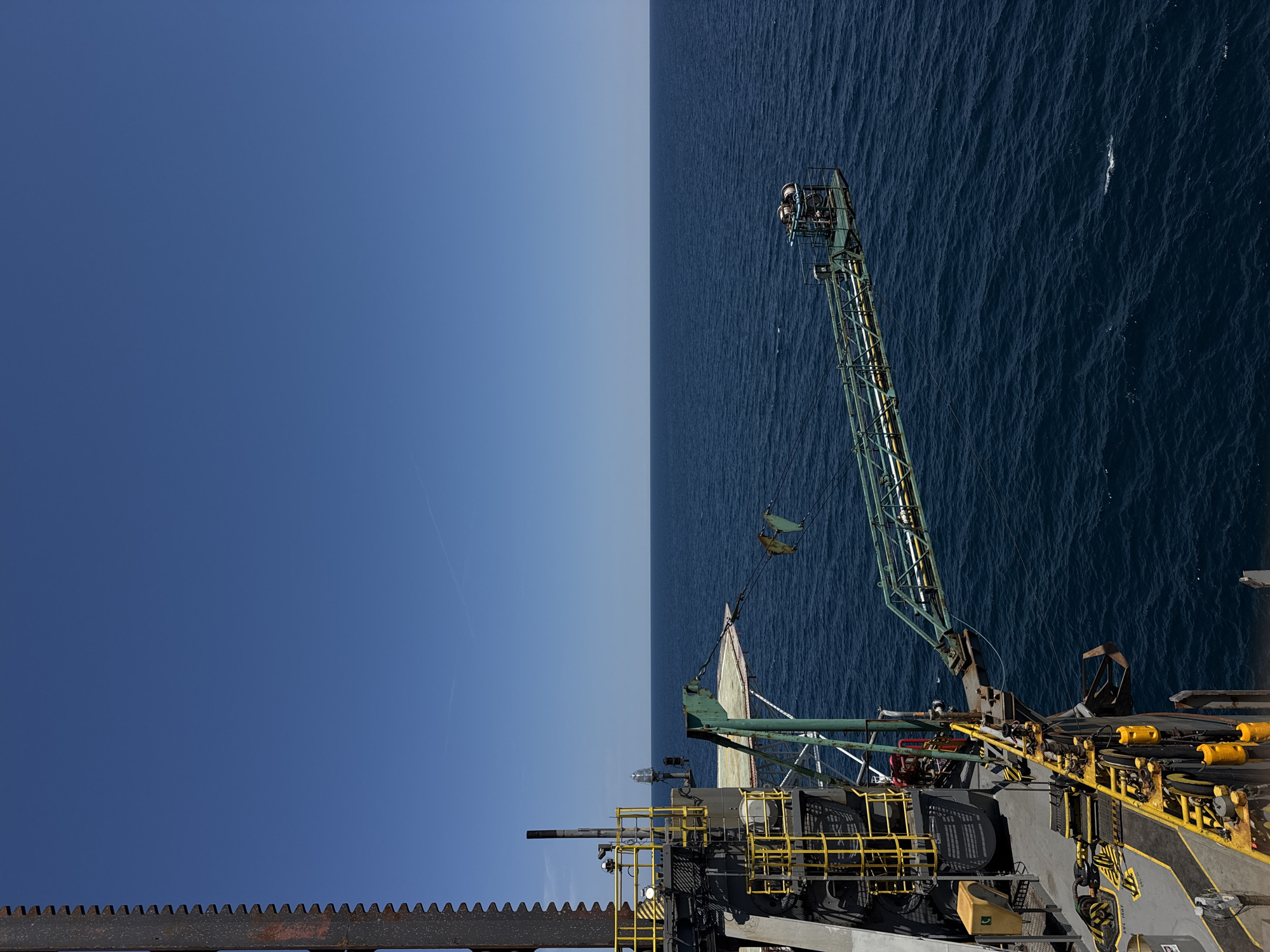 The image shows a view from an industrial platform or ship on the open ocean, under a clear blue sky. In the foreground, a large, dark, toothed structure, possibly part of a drilling rig or construction equipment, dominates the left side of the frame. Yellow safety railings and industrial equipment are visible on the platform. Extending from the platform over the water is a green, gantry-like structure with pipes and machinery at its end. The dark blue ocean stretches out to the horizon, with small ripples on its surface. There are no people or animals visible in the image. The scene appears to be in an industrial maritime setting, not in a city.