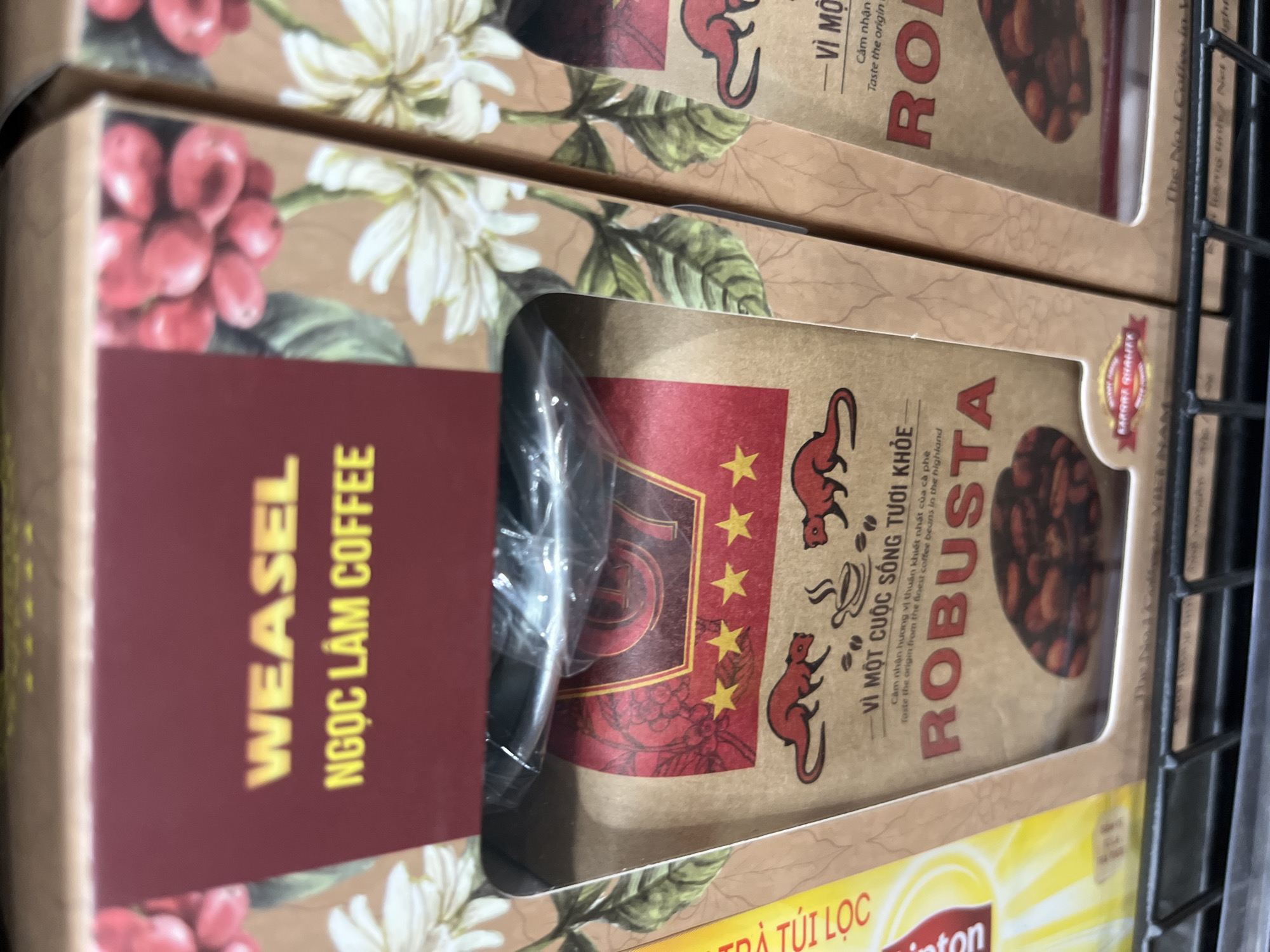 The image shows a close-up of two boxes of "WEASEL NGỌC LÂM COFFEE ROBUSTA" coffee. The boxes are brown with red accents and feature illustrations of coffee cherries and flowers. The left box has a clear plastic window revealing a coffee bag inside, which is also brown with red graphics. The text "VI MỘT CUỘC SỐNG TƯƠI KHỎE" and "ROBUSTA" are prominently displayed. The boxes are stacked on a black metal shelf, suggesting they are on display in a retail environment, possibly a market or a store in Nha Trang Ward, Vietnam. There are no people visible in the image.