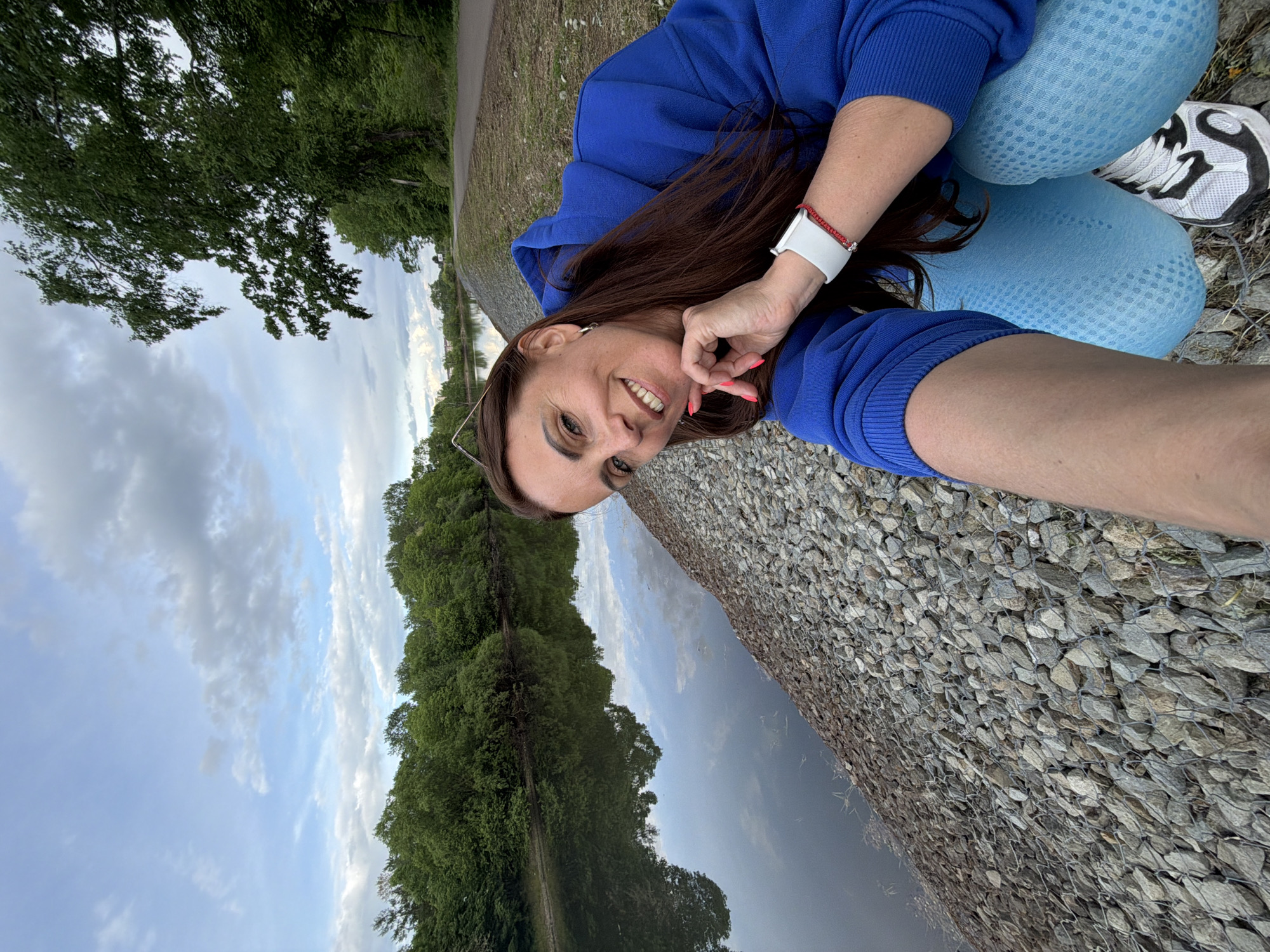 A woman is captured in a selfie outdoors, displaying a wide smile directly at the camera. She has long, dark brown hair and is wearing a royal blue hooded sweatshirt and light blue patterned leggings. Her left arm is raised, with her hand near her chin in a relaxed pose, revealing a white smartwatch and a red beaded bracelet on her wrist, and light pink nail polish. Sunglasses are perched on her head. Her left foot, clad in a white and black athletic shoe, is also visible.
The scene depicts a natural setting, likely a park or lakeside area. To the woman's right, there is a gabion wall, constructed from rocks held within wire mesh. Beyond this wall lies a calm body of water, possibly a lake or a wide river, which reflects the sky. A dense line of lush green trees lines the far bank of the water, extending into the distance. To the woman's left, a paved path is visible, bordered by more greenery. The sky above is partially cloudy, suggesting a daytime setting with diffused light, perhaps late afternoon or early morning. The overall atmosphere is serene and pleasant, indicative of an outing in nature. The location is in Birobidzhan, Russia. No visible text is present in the image.