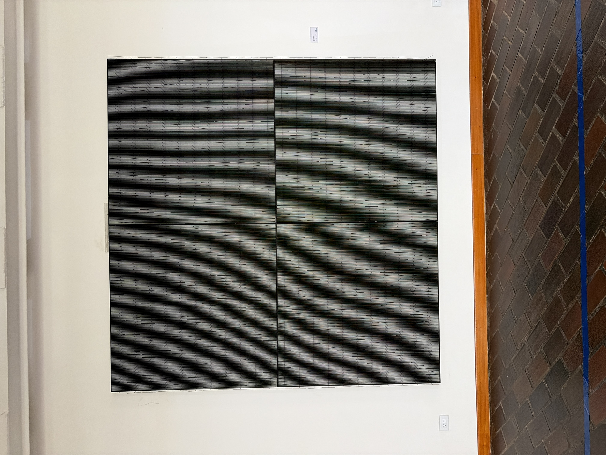 The image shows a large, square artwork composed of four panels with horizontal lines. The artwork is mounted on a white wall. There's a light switch or outlet below the right bottom panel and a small white label above the top center point of the artwork. To the left is a white wall with a white vertical element. To the right, there is a brick wall or floor with a vertical blue line taped. It is not possible to determine the location in Ciudad Bolivar, Venezuela, from the given image. No people are visible in the image.