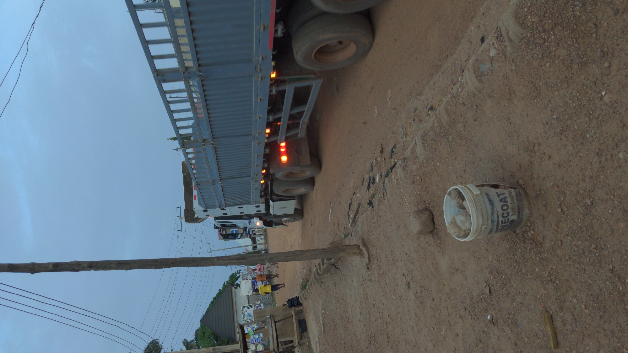 The image depicts a street scene, possibly in a commercial area. A large truck is parked along a dirt road. A utility pole with visible wires runs along the side of the road. A discarded NECOAT paint bucket sits on the ground near the truck. In the background, there are buildings, people, and various objects, suggesting a lively, populated area. The image suggests a roadside scene with evidence of human activity and commerce. No specific details confirm the location is in Masaka, Nigeria.