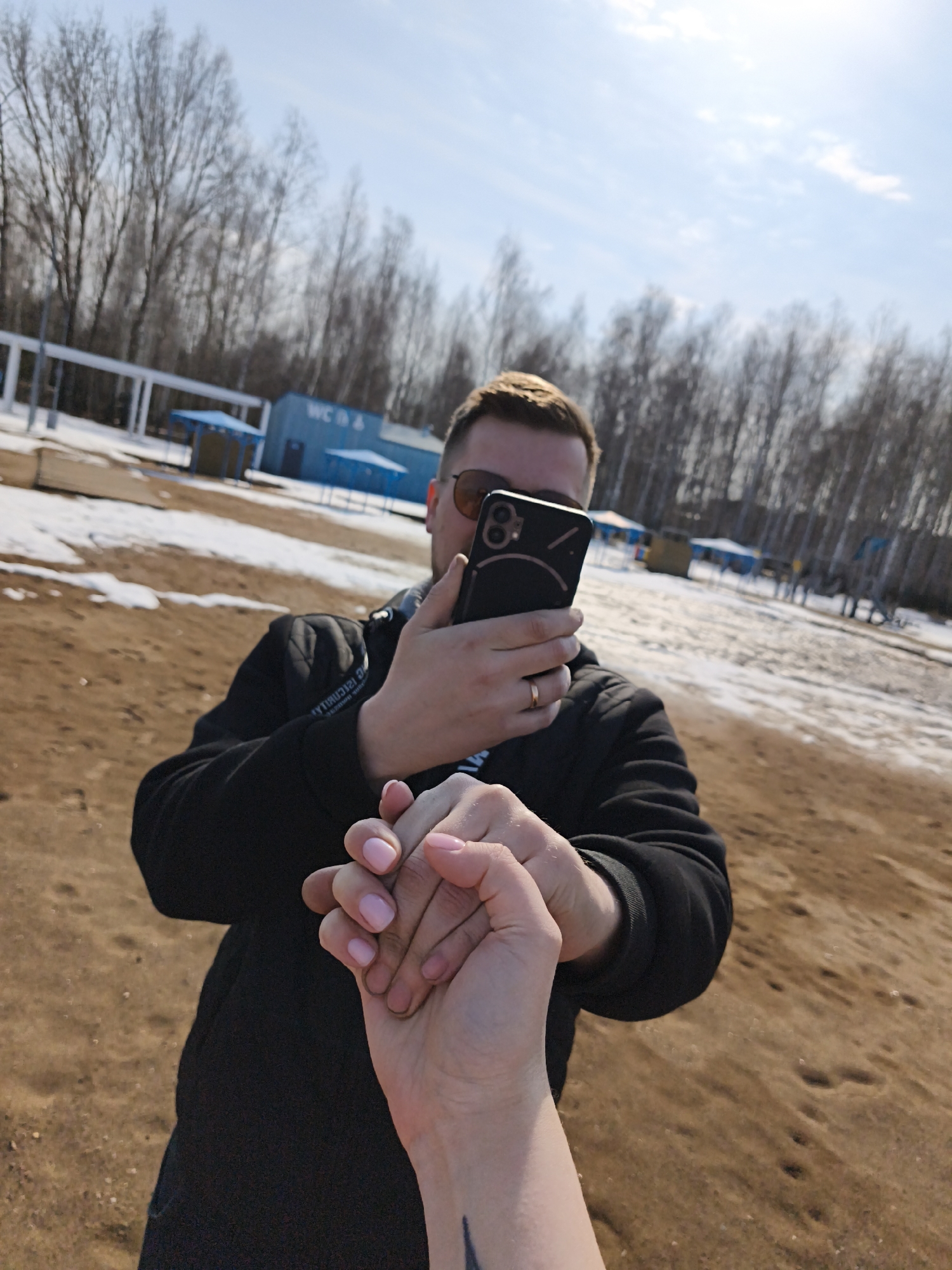 A medium shot depicts a man holding a mobile phone while grasping a hand in front of him. The man, wearing sunglasses and a black jacket, appears to be taking a photo with his phone. He has a gold ring on his left ring finger. The hand he is holding, presumably belonging to another person, has pink nail polish and a small tattoo is visible on the wrist. The setting appears to be outdoors in a location with a sandy surface and patches of snow. In the background, there are bare trees and a building with "WC" visible on it, along with several blue structures. The sky is light blue with some wispy clouds. The location appears to be in the city of Unknown, Unknown.
