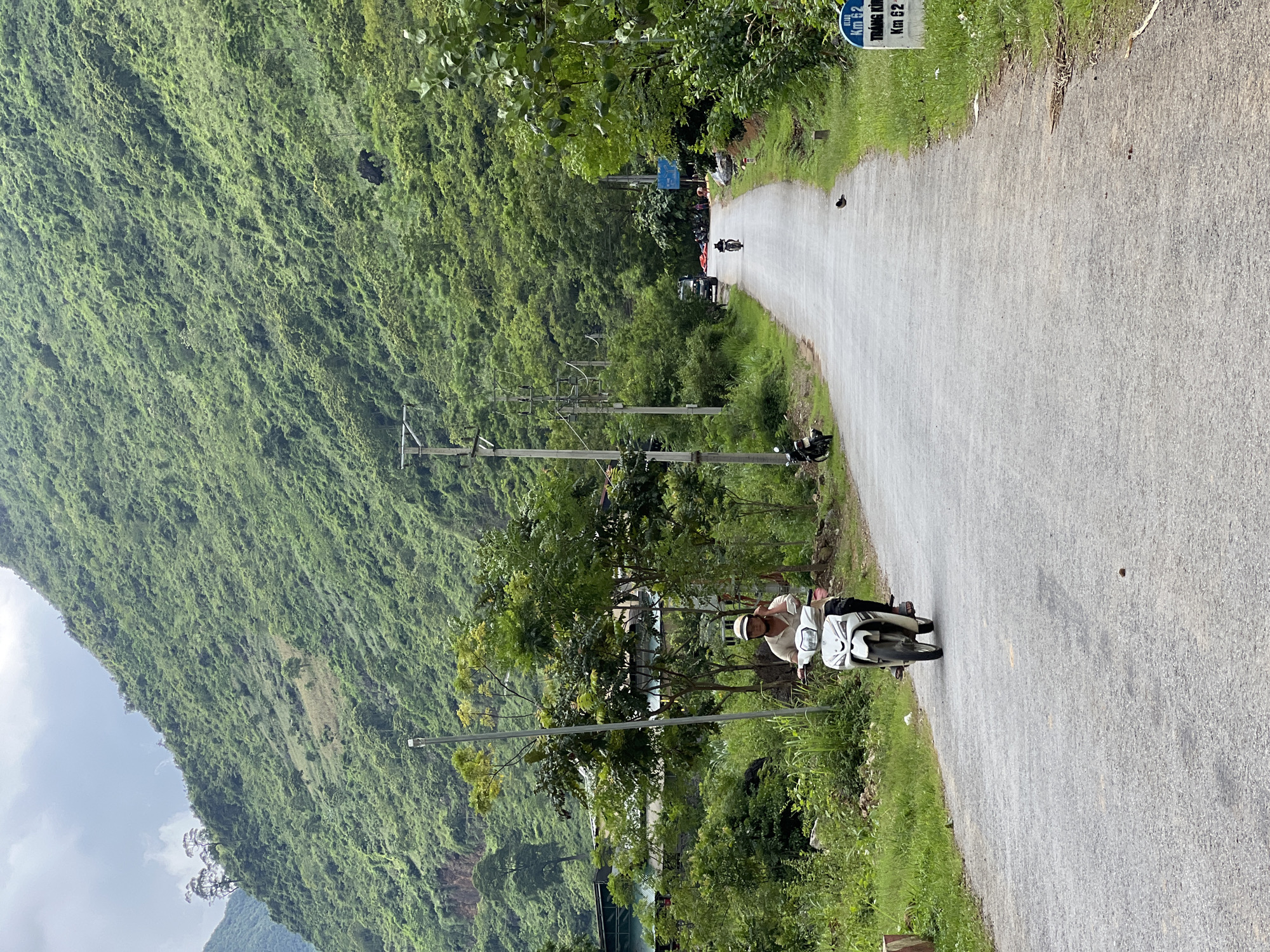A person is riding a white scooter down a paved road. The road is flanked by lush green vegetation and a steep, heavily forested mountain on the left. The sky is overcast. The road curves to the right in the distance where several other motorcycles and a few people are visible. There are utility poles lining the left side of the road. A white sign with blue text, reading "KM 62 TRANG KIM KM 62," is visible on the right side of the road. The setting appears to be a rural mountainous area in Vietnam, consistent with locations like Cán Tỷ Commune.