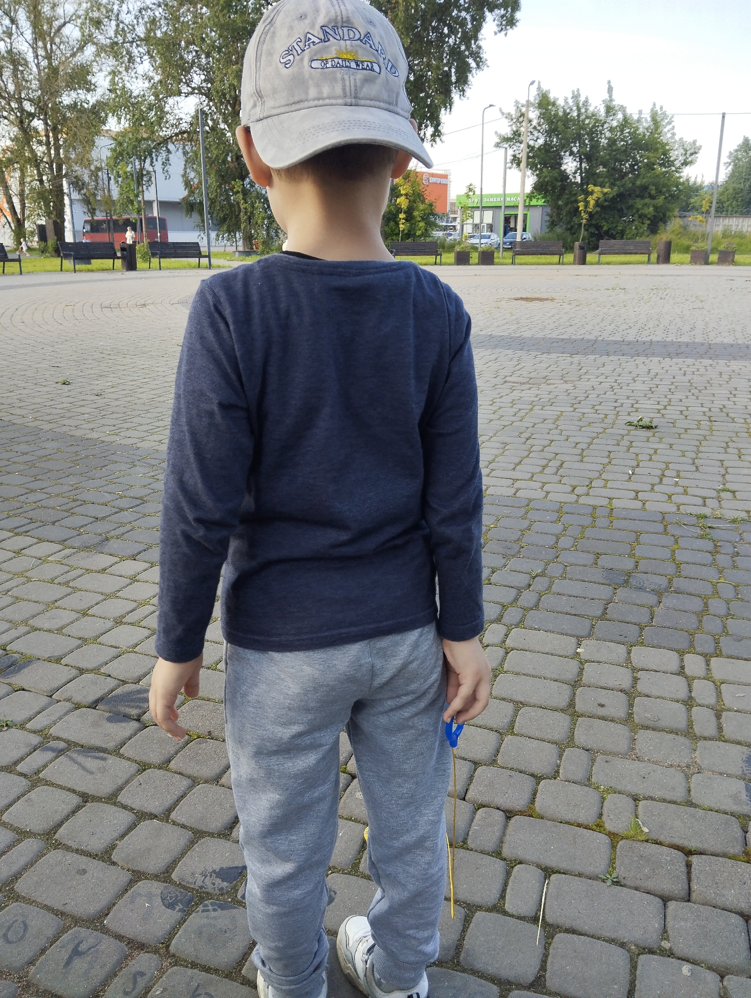 A young boy, viewed from behind, stands on a cobblestone plaza in Kingisepp, Russia. He wears a grey baseball cap, a navy blue long-sleeved shirt, and light grey sweatpants. His right hand holds a blue and yellow string toy. In the background, trees line a street with buildings and parked vehicles, including a red bus. Several park benches are visible on the plaza. The sky is overcast.