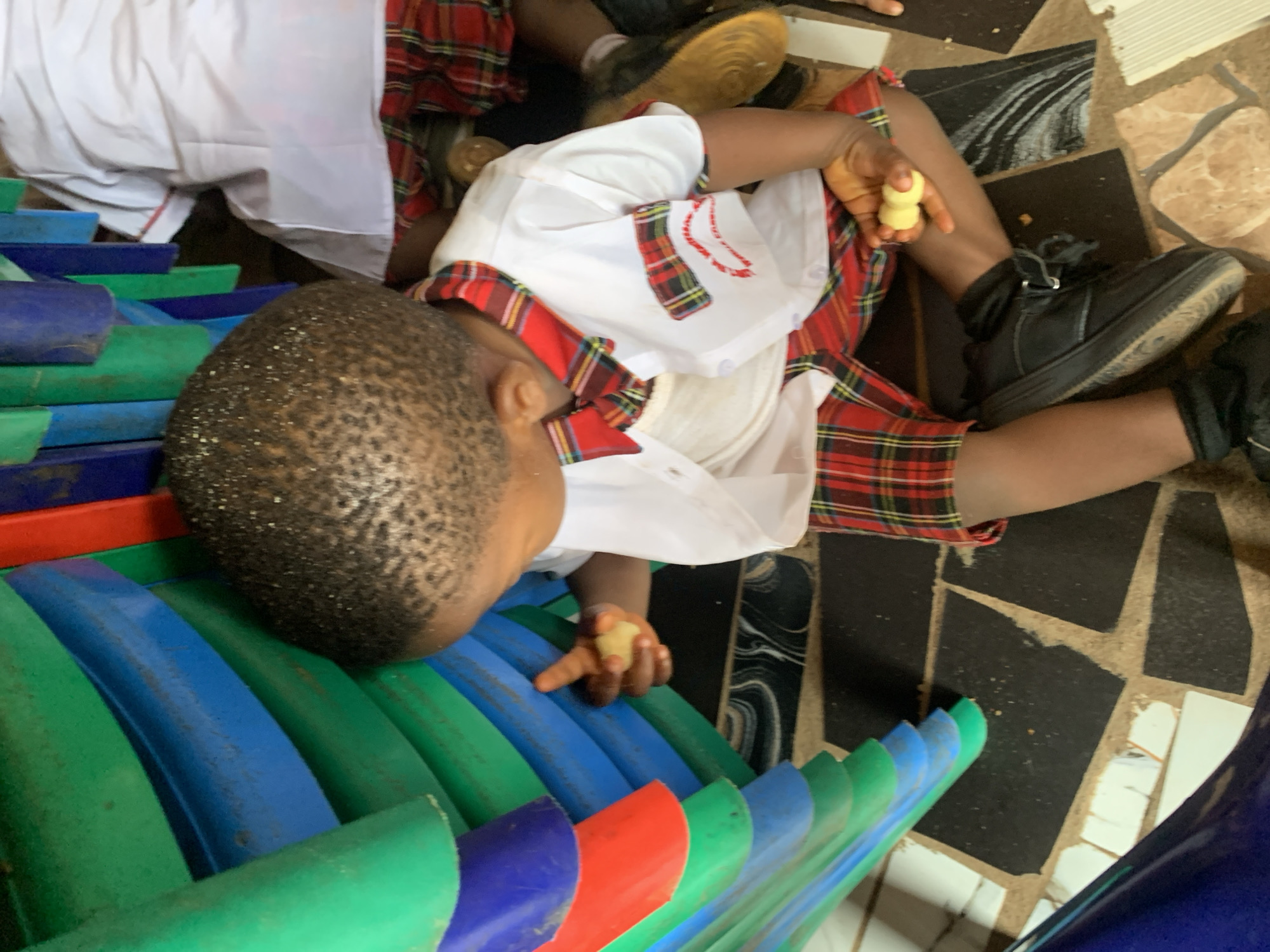 A child in a school uniform with a red and black plaid skirt and white shirt with red plaid trim lies on a floor with patterned tiles in Ukwu-Abwa, Nigeria. The child rests their head on a stack of plastic chairs in green, blue, and red, holding a small object in each hand. One hand is on the chairs and the other is near their leg. The child is wearing black shoes and appears to be eating. Another child in a similar uniform and white pants is partially visible in the background.