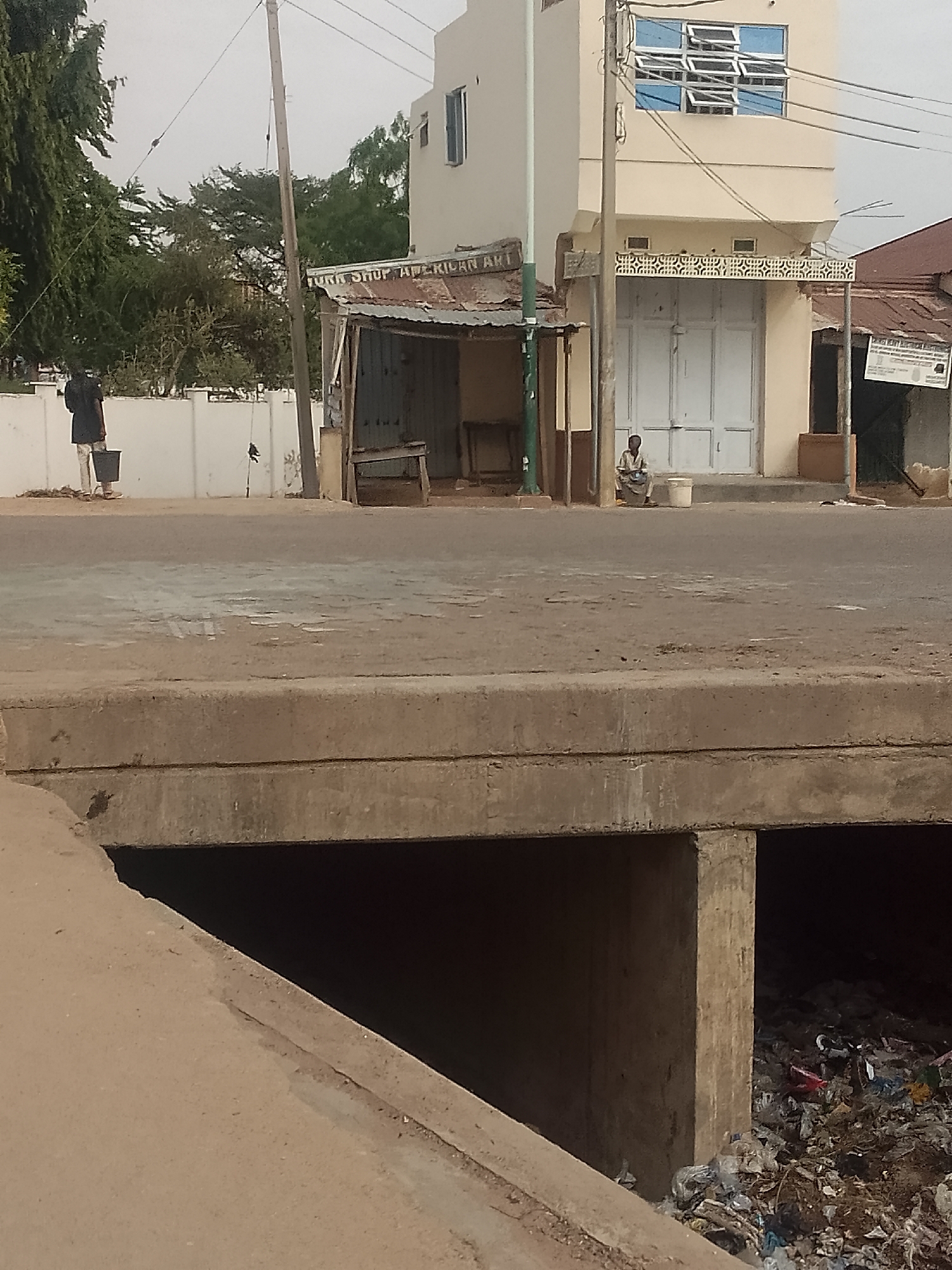 The image shows a street scene in Pantame, Nigeria. In the foreground is a concrete drainage culvert filled with garbage. Across the street, a man in a dark shirt and light pants is carrying a bucket. Further down the street, a person is sitting on a bucket in front of a building with closed white doors and a sign with decorative scrollwork above the door. A small shop with the words "TURK SHOP AMERICAN ART" is to the left of this building. Trash is piled in the drainage culvert. A utility pole is beside the shop.