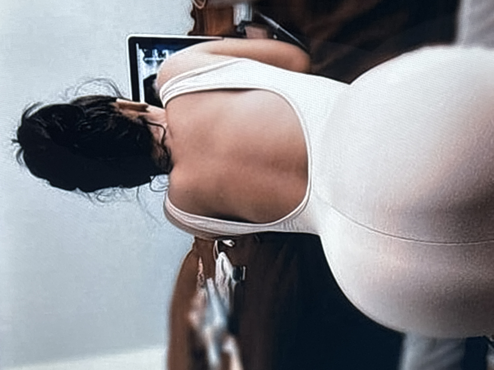 A woman with dark hair pulled into a bun is wearing a white bodysuit and standing indoors. She is looking at a laptop screen, which is slightly out of focus. Another person is partially visible behind her, wearing brown clothing. The background is a blurry white wall. The location is not identifiable as Saint Petersburg, Russia based on the image.