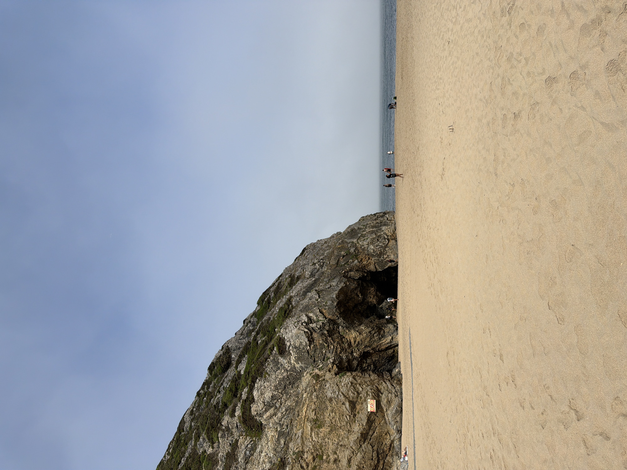 A wide shot captures a sandy beach leading to a large rock formation with a cave opening. The beach is mostly empty, with a few scattered individuals. To the left, a person is lying on the sand. Near the cave entrance, several people are visible, some appearing to enter or exit the cave. The ocean stretches across the background, meeting a hazy sky. A warning sign is affixed to the rock face to the left of the cave. The overall atmosphere is tranquil and natural, consistent with Praia da Adraga in Colares, Portugal.