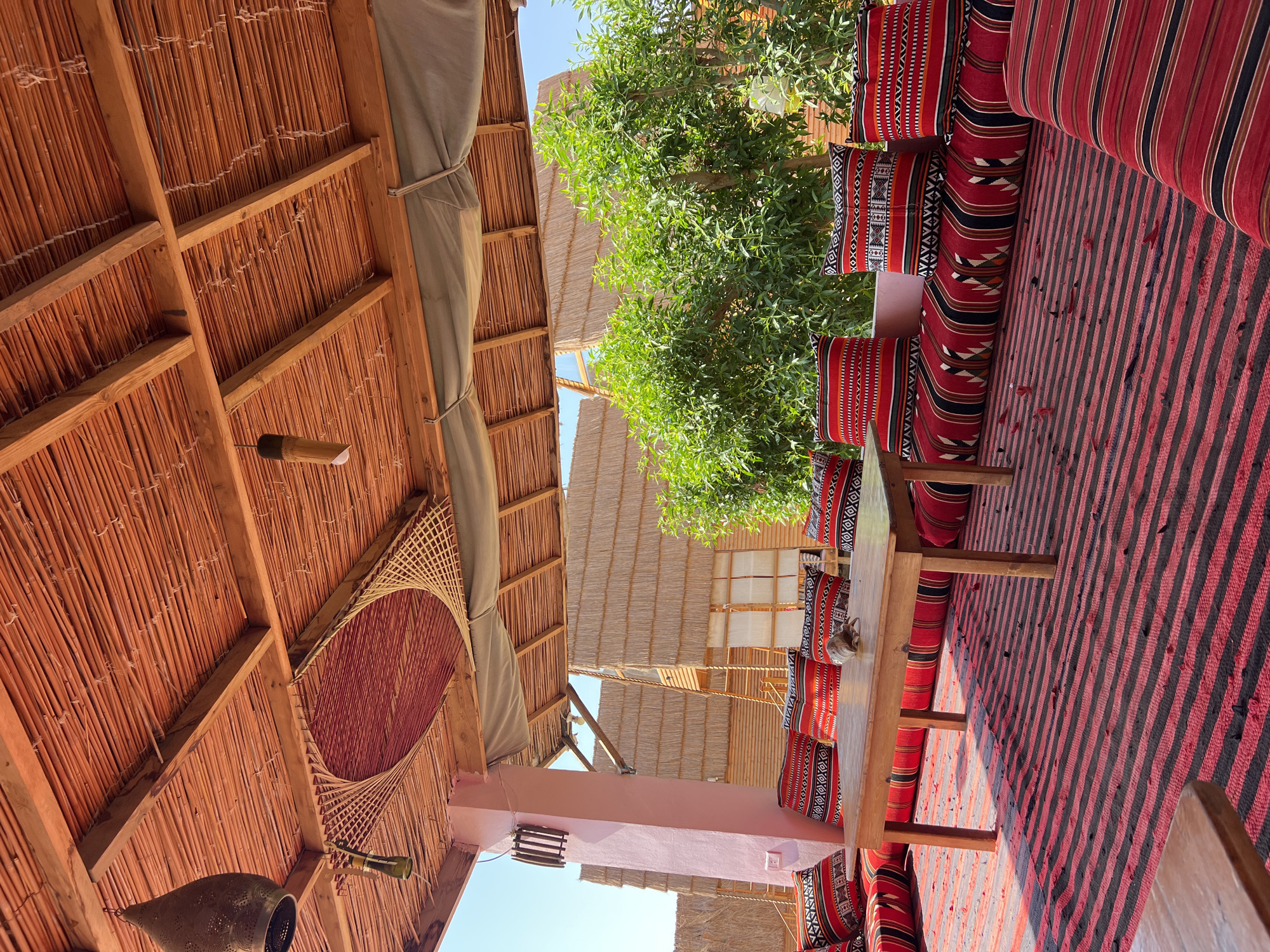 This image depicts an outdoor seating area with a traditional, rustic aesthetic, likely located in Nuweiba, Egypt, given the architectural style and vegetation. The scene is viewed from a low angle, looking towards a covered seating arrangement.
The ceiling is constructed from woven reeds or similar plant material, supported by wooden beams. Two decorative light fixtures are suspended from the ceiling: one is a large, metallic, lantern-like object, and the other is a smaller, cylindrical bamboo fixture. A circular, woven decorative element is also attached to the ceiling.
The seating area features a long, low sofa upholstered with red and black striped fabric, piled with numerous square cushions. These cushions are adorned with intricate geometric patterns in red, black, white, and yellow. A simple, light-wood coffee table with four legs sits in front of the sofa. On the table, a single shell or similar artifact is placed.
In the foreground, a large rug with bold red and black horizontal stripes covers the ground. Scattered across the rug are small, red flower petals or leaves. Another wooden table is partially visible in the lower left corner.
Behind the seating area, lush green trees with dense foliage are visible. In the background, several small structures with thatched roofs and wooden walls are present, suggesting a resort or a cluster of cabins. The sky is clear and bright blue. The overall impression is one of a relaxed, bohemian outdoor lounge area.
