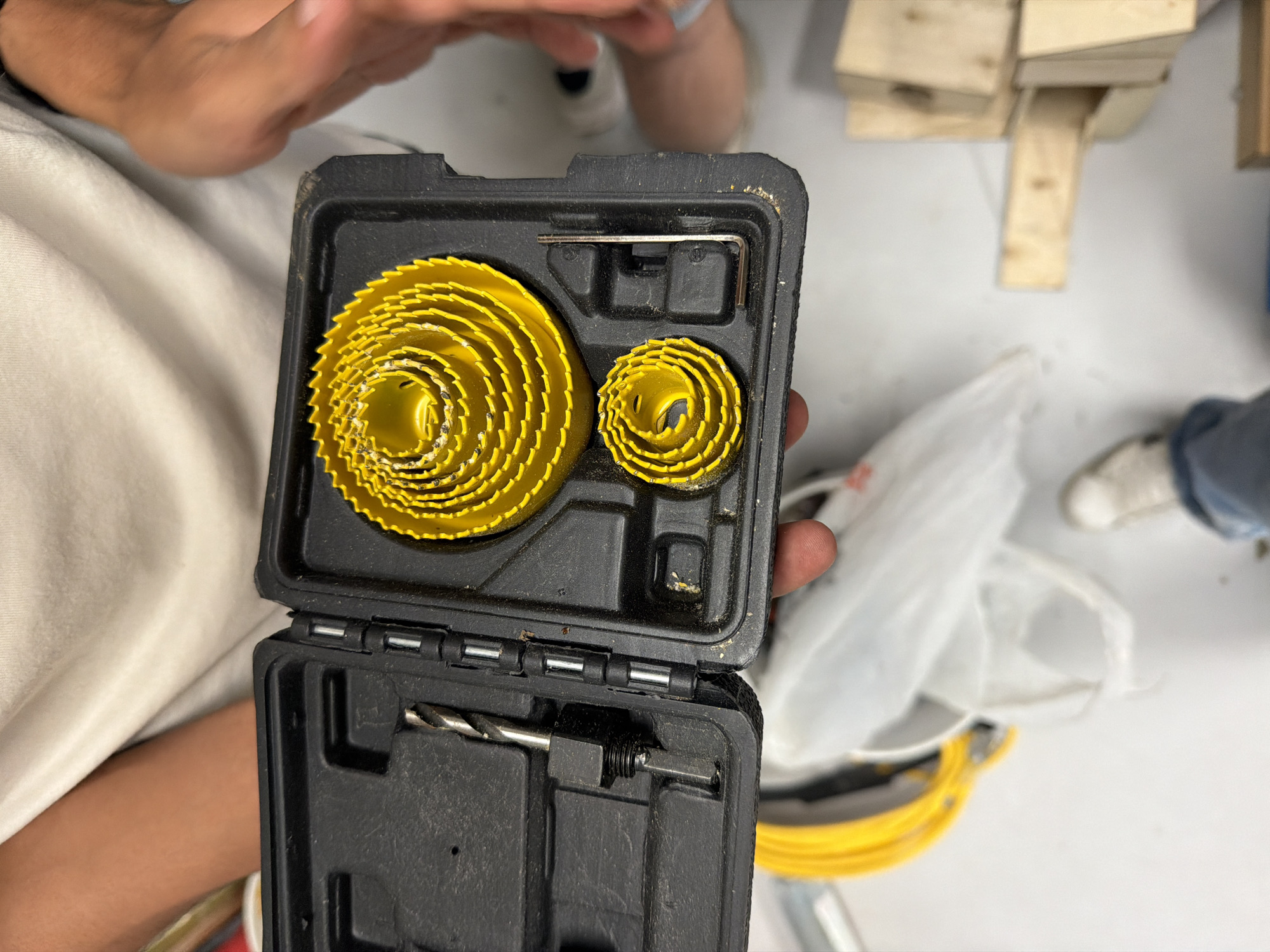 The image shows a close-up, top-down view of an open black plastic tool case, held by hands, revealing its contents. The primary objects within the case are a set of yellow hole saw blades, organized by size. A larger stack of progressively smaller saws is on the left, and a smaller stack is on the right, both neatly nestled in custom-molded compartments. Also visible in the top section of the case is a small, L-shaped hex key (Allen wrench). In the lower half of the opened case, a silver drill bit, which appears to be an arbor for attaching the hole saws, is secured in its own slot.
The subjects include a person's hands holding the tool case, with fingers partially visible at the edges of the frame. In the background, other parts of people, including an arm and legs with clothing (one wearing a white shoe), are vaguely discernible, suggesting multiple individuals are in the vicinity.
The setting appears to be an indoor workshop or garage, indicated by the utilitarian nature of the tools and the background elements. These elements include several light-colored wooden blocks or scraps stacked on a surface in the upper right portion of the background, and a white plastic bag along with some yellow coiled cords and a white shoe on the floor or a lower surface to the right. The floor or main surface seems to be light-colored.
The activity depicted is the presentation or viewing of the hole saw kit, as it is held open for inspection. The tools and case show some signs of use, with minor dust or debris visible. No specific time of day, weather, or discernible emotions are evident in the image. There is no visible text on the tools or the case itself.