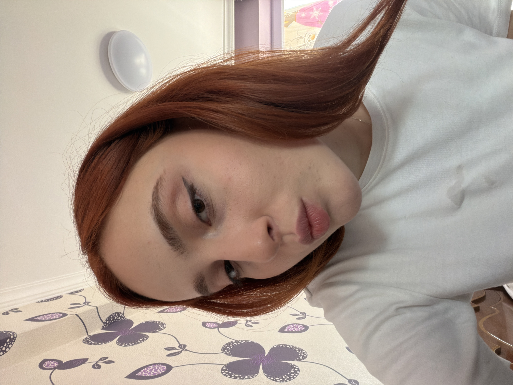 A selfie of a fair-skinned person with auburn hair wearing a white top. The person's face is angled towards the camera with a focus on their eyes, nose, and lips. The background includes a white wall with a circular light fixture, wallpaper with purple floral patterns, and a glimpse of a window with light-colored curtains. The location is Tbilisi, Georgia, based on the architectural style of the room.
