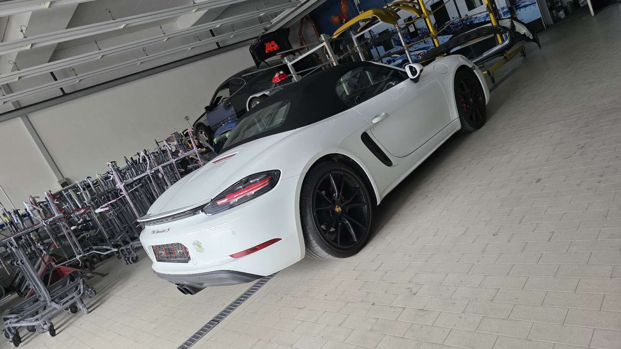 A white Porsche 718 Spyder S convertible is parked in what appears to be a workshop or garage. The car has a black soft top and black wheels with red brake calipers. In the background, there are several other vehicles, including a dark blue car and a black car. Racks filled with what appear to be car parts or equipment are also visible, as are some shopping carts. The floor is tiled with a light grey pattern. There are no people visible in the image.