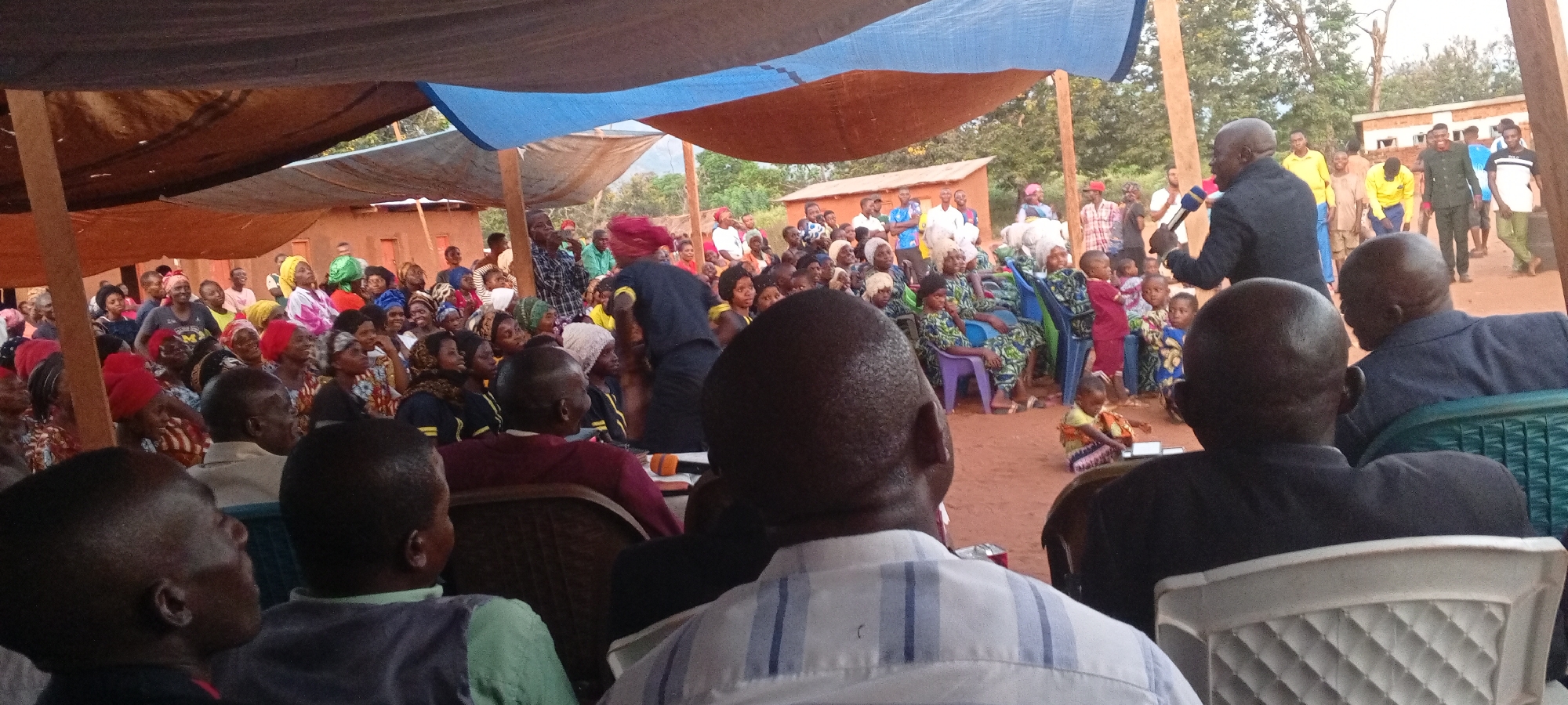 A crowded outdoor gathering is taking place in Kalemie, Democratic Republic of the Congo. An African man in a dark suit is standing and speaking into a microphone, addressing the crowd. People of all ages, including children, are seated or standing, watching and listening to the speaker. The crowd is predominantly black. Many are wearing colorful clothing, and some women have head coverings.
A temporary shelter has been constructed with fabric tarps suspended overhead. A few simple buildings and trees are visible in the background, suggesting a rural or semi-rural setting. The ground appears to be reddish-brown dirt.