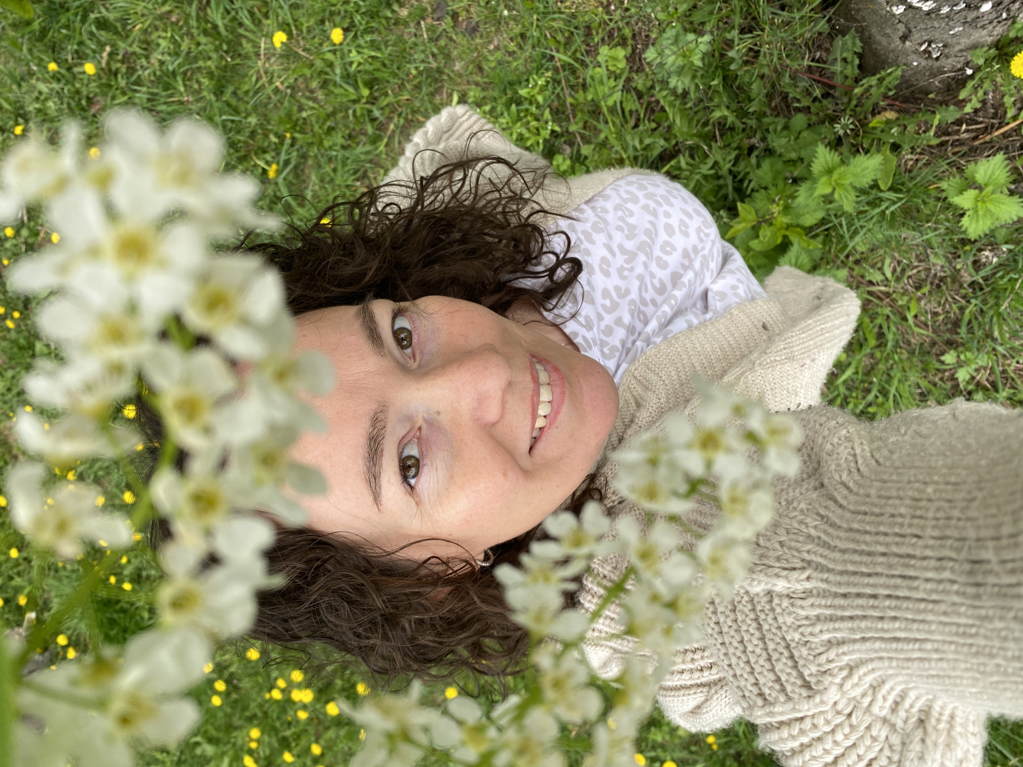 A woman lies on her back in grassy terrain dotted with dandelions, her face framed by blooming white flowers. She is fair-skinned with curly brown hair and brown eyes. She wears a light-colored knit sweater over a top with a beige and white patterned design. The background consists of a green grassy field with a few patches of small green plants, some of which appear to be nettles, and what looks like the edge of a tree trunk in the upper right corner. The exact location in Ольховка, Russia, cannot be determined from the image.