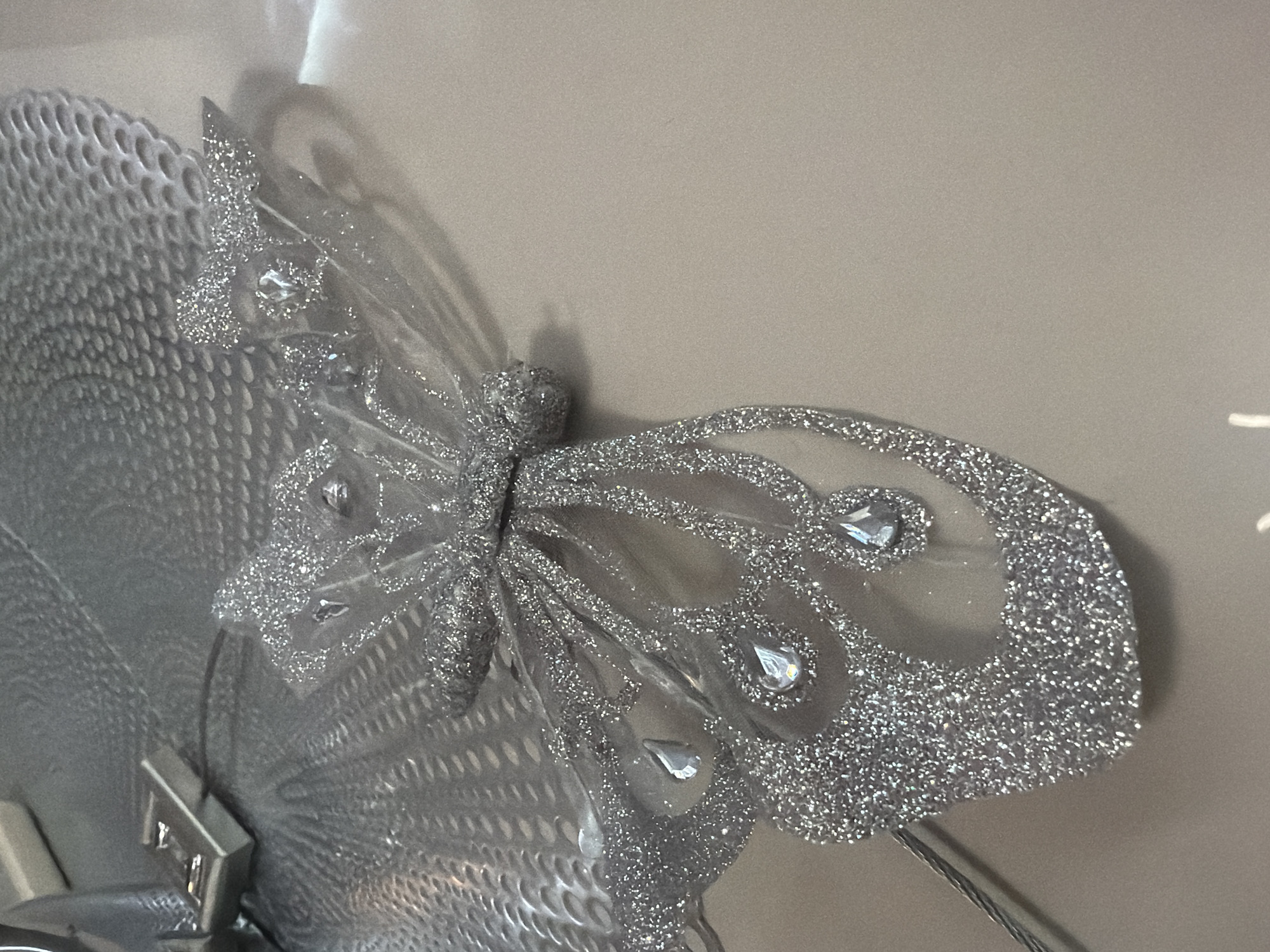 A close-up shot reveals a decorative, silver, glitter-covered butterfly. The butterfly is adorned with clear, teardrop-shaped jewels. Its wings are partially transparent, and the edges are heavily coated in glitter. The butterfly is attached to a textured, silver, perforated surface. A metallic, possibly chrome, object is visible near the attachment point. The background is a plain, light beige wall.