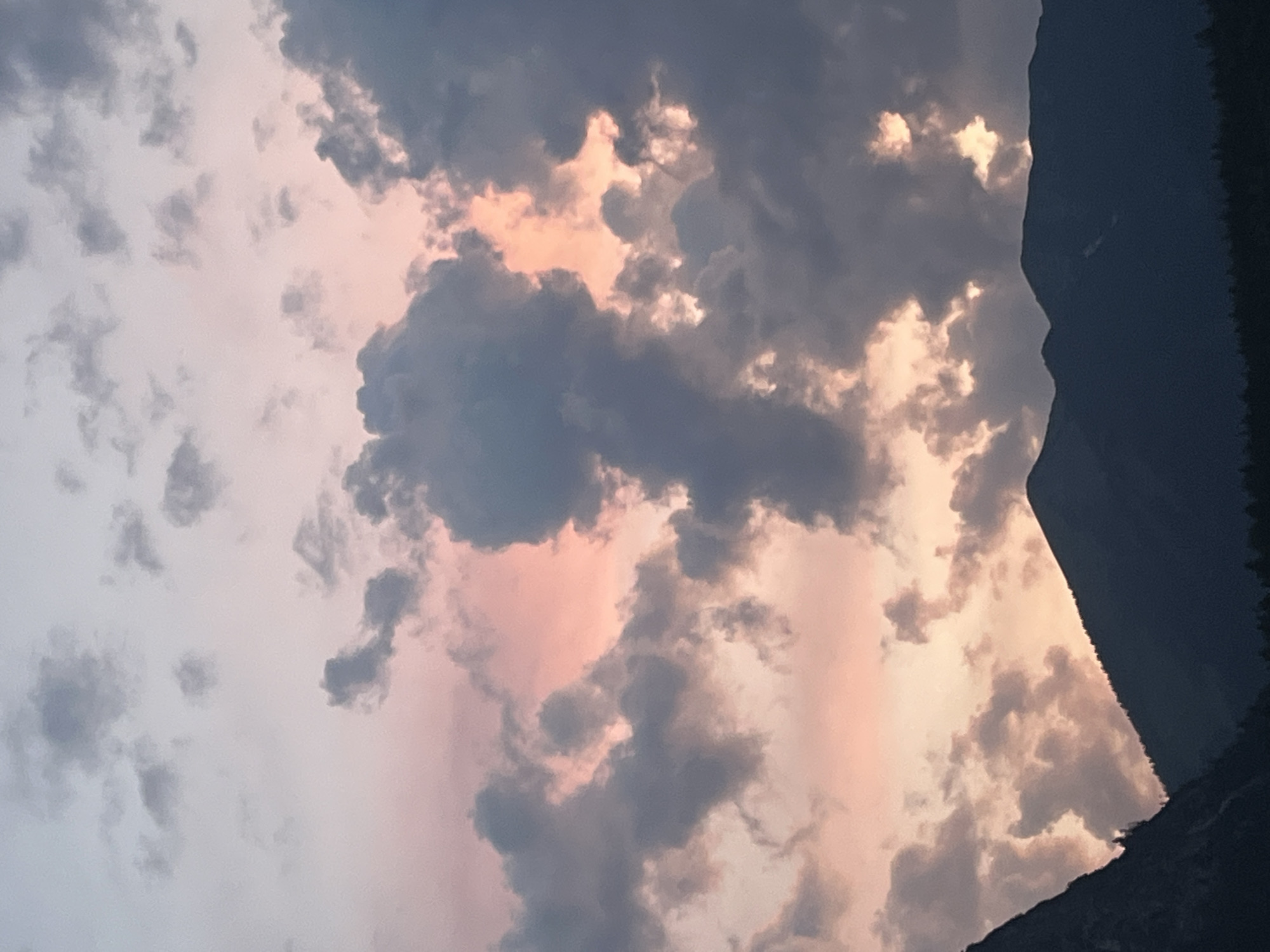 This is a vertical, medium-shot, eye-level photograph of a mountain range in Garmisch-Partenkirchen, Germany, against a dramatic, cloudy sunset sky. The foreground is dominated by the dark silhouettes of two jagged mountain peaks, with a dense line of trees at their base. The sky above is a canvas of pinks, oranges, and grays, with thick, dark clouds obscuring much of the vibrant sunset colors. Some patches of lighter pink and orange peek through the clouds, creating a sense of depth and intensity. The overall mood is serene and majestic. There are no people or animals visible in the frame.