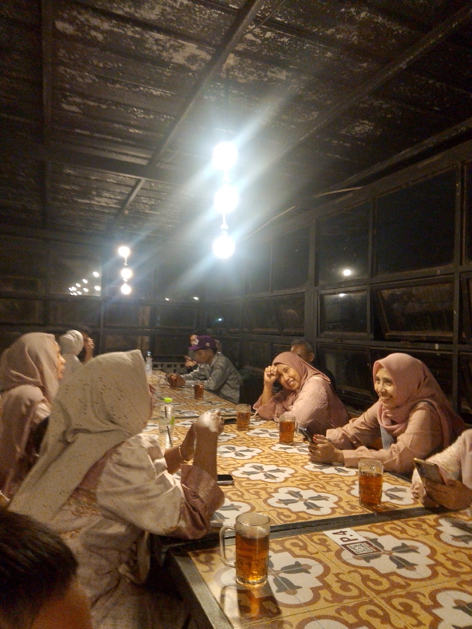 The image depicts an indoor gathering of people, likely in Tasikmalaya, Indonesia, as suggested by the provided information. The scene appears to be a casual dining or social event taking place under a rustic, exposed ceiling with multiple hanging light bulbs. A group of individuals, predominantly women wearing hijabs, are seated around a patterned table. The table is set with glasses of beverages, and one person is holding a mobile phone. The lighting is dim, with the hanging bulbs providing the primary illumination, creating a warm, intimate atmosphere. The background features large windows, suggesting the setting might be a restaurant or an outdoor-covered area. The overall impression is of a social gathering, possibly a meal or a relaxed conversation, in a dimly lit, atmospheric venue.