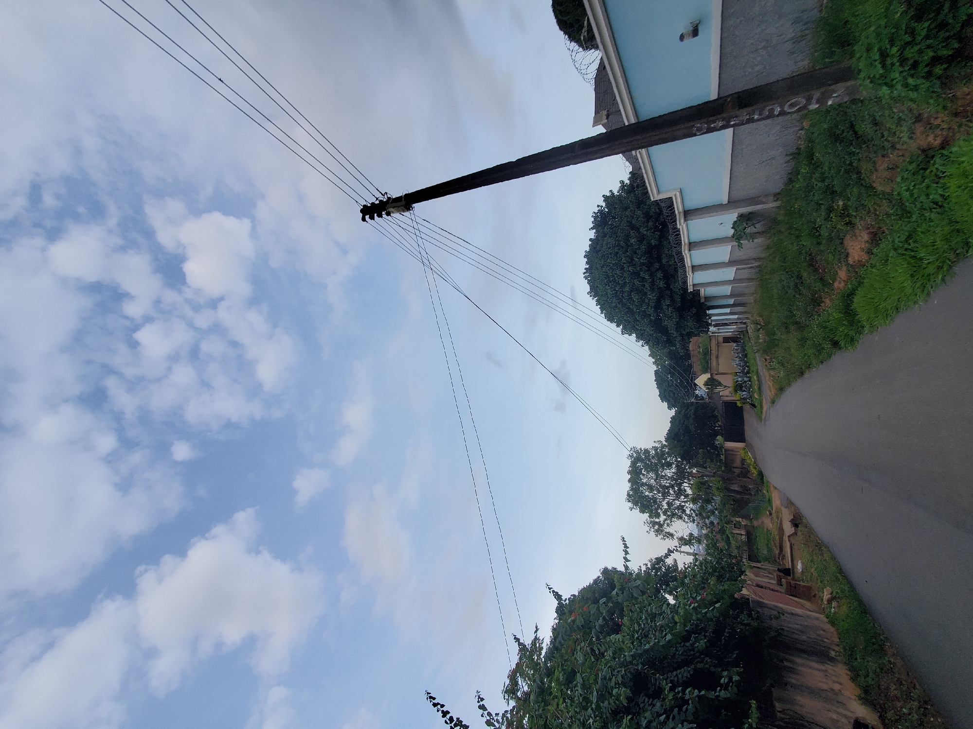 This image depicts a street scene in Jos, Nigeria, likely during the early morning or late afternoon given the soft lighting and cloudy sky. The foreground features an asphalt road curving gently into the distance. To the left of the road, a dense wall of lush green foliage with some red flowers spills over a rough, aged concrete wall. Further down the road, trees of varying heights and densities are visible, interspersed with some buildings.
On the right side of the road, a tall, weathered wooden utility pole stands prominently, supporting multiple dark power lines that stretch across the frame. Adjacent to the pole is a light blue wall topped with grey concrete and barbed wire, suggesting a secured property. Beyond this wall, more trees and structures are visible. The sky is a pale blue, scattered with fluffy white clouds. The overall impression is of a semi-urban or residential area with a mix of natural greenery and man-made infrastructure.