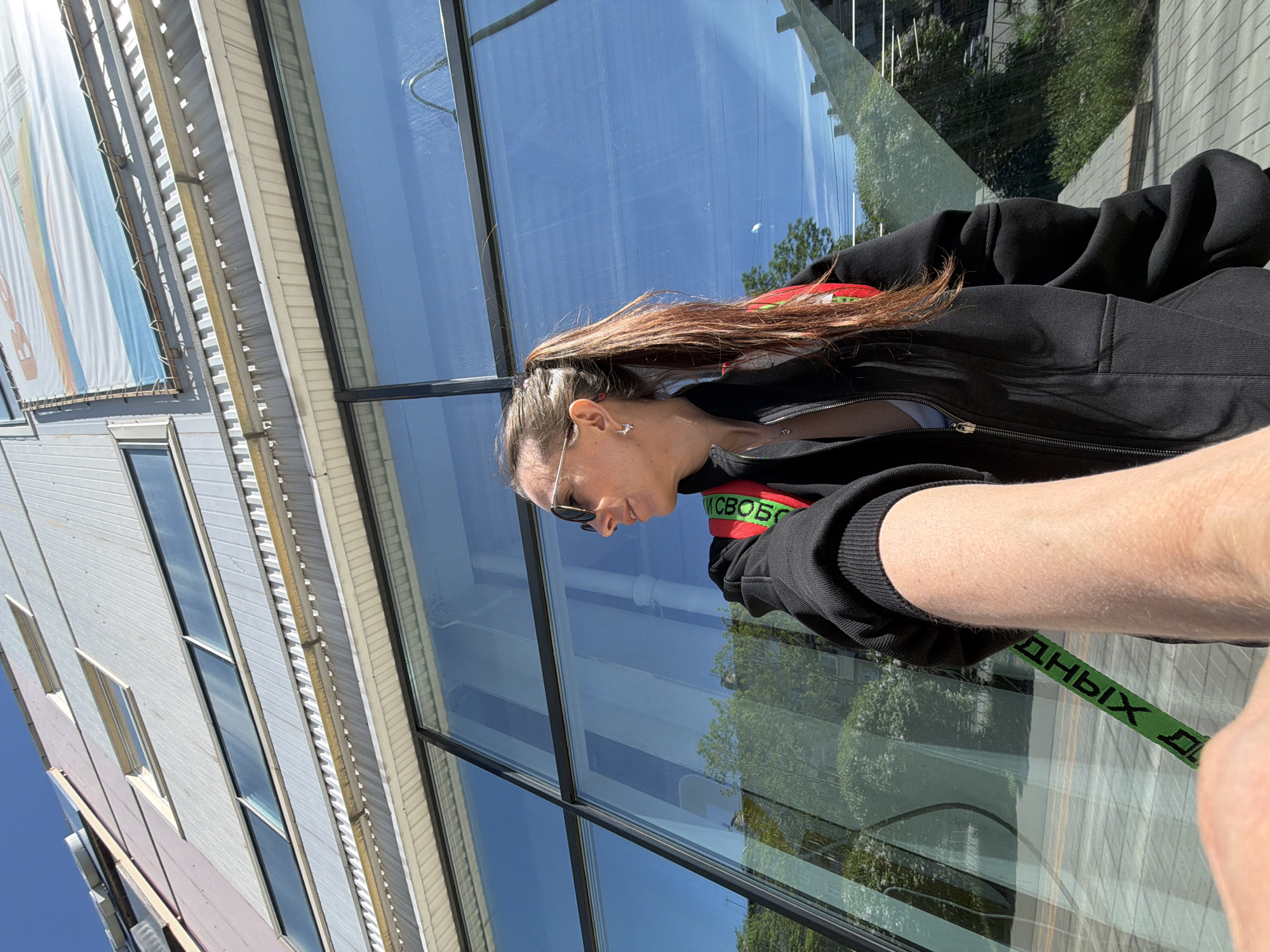 A medium shot captures a woman standing next to a modern building with large, reflective windows. The building, possibly located in Birobidzhan, Russia, features a gray façade with a billboard on its left side. The woman is wearing sunglasses and a black jacket with a green and red strap across her shoulder, partially obscuring the view of the building and reflecting the surroundings in the glass.