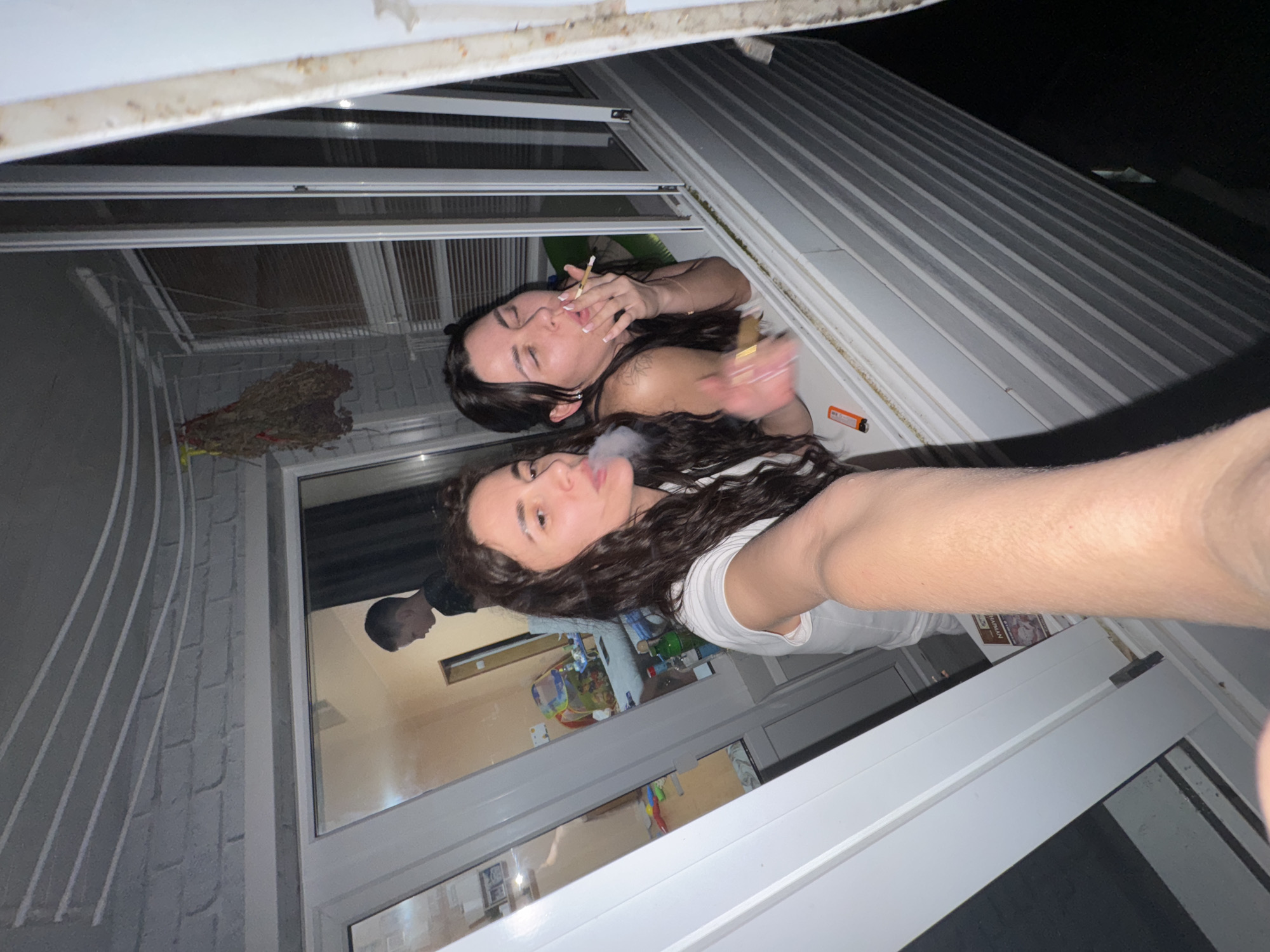 Two women are positioned in a window, one behind the other, and both are smoking. The woman in the foreground has dark curly hair, is wearing a white t-shirt, and is exhaling smoke. The woman in the background has dark hair, is holding a cigarette to her lips, and is topless. Part of a man's face is visible in the reflection of the window behind the women. The environment is part of a building, with a white frame around the window. A lighter is visible near the woman smoking in the background. The location is Chaikovsky, Russia.