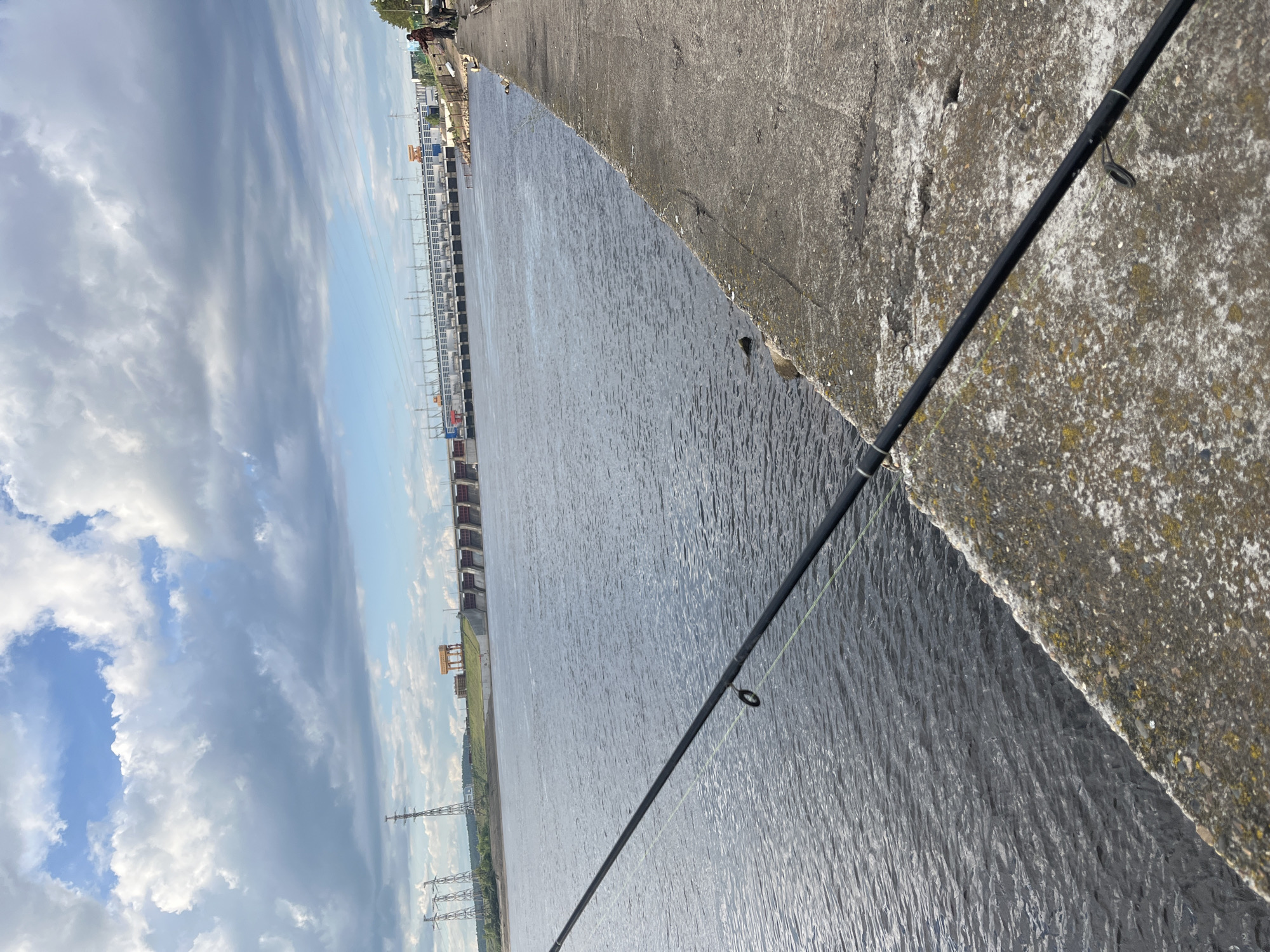A fishing rod extends from a concrete embankment, casting a line into the waters of the Kama River in Chaikovsky, Russia. In the background is a dam complex. Above, a cloudy sky dominates the scene. High-voltage power lines stretch away along the riverbank.