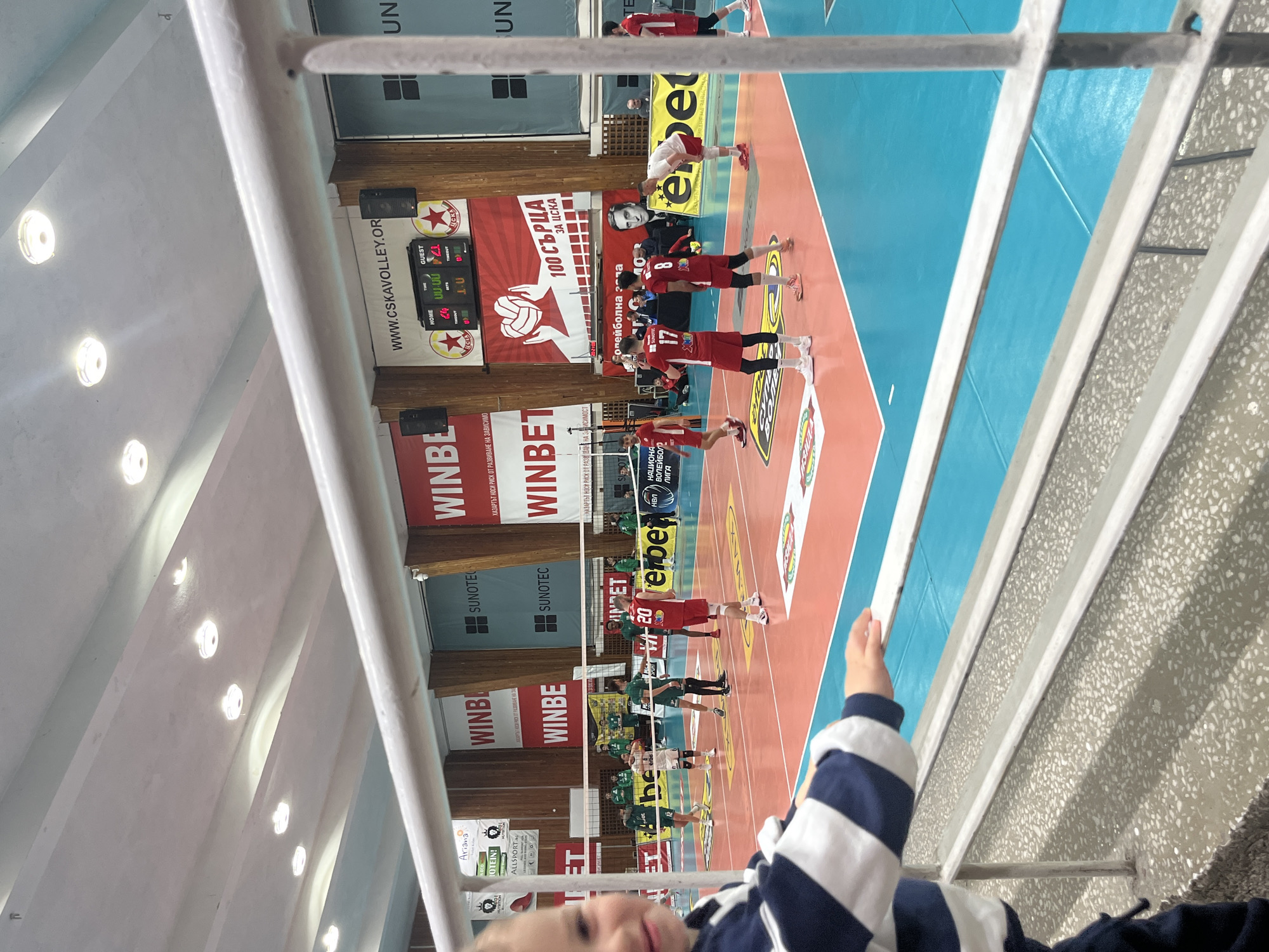 An indoor volleyball game is underway in a sports hall, specifically a CSKA facility in Sofia, Bulgaria, identifiable by the "WWW.CSKAVOLLEY.ORG" website, "ЦСКА" logos on the scoreboard, and "100 СЪРЦА ЗА ЦСКА" (100 Hearts for CSKA) banner.
In the immediate foreground, a young child, partially visible from the chest up, wears a navy blue and white striped long-sleeved shirt and dark pants. The child's left hand rests on a white metal railing, and the face is turned slightly towards the viewer with a subtle smile, appearing blurred.
Beyond the railing, a multi-colored volleyball court is visible, featuring a central orange-red playing area bordered by light blue. White lines delineate court boundaries. Two teams of male players are on the court. One team wears red jerseys with white numbers; prominent players include number 17 (wearing black arm sleeves and leggings) and number 20 (also in black leggings). The opposing team wears green and white jerseys; player 12 in a green jersey is visible. Players are in various stances, indicating active play or a pause between points. A white net spans the center of the court.
The background consists of the hall's walls adorned with large advertising banners for "WINBET," "SUNOTEC," and "erbet," along with a banner featuring a portrait of a man. A central electronic scoreboard displays "HOME: 0," "GUEST: 21," and "SETS: 1:0," with "ЦСКА" logos. The ceiling is grey concrete with exposed beams and recessed circular light fixtures, providing bright illumination. Black speakers are mounted on the wall near the scoreboard. The spectator area in the foreground features a grey, speckled concrete or terrazzo floor.