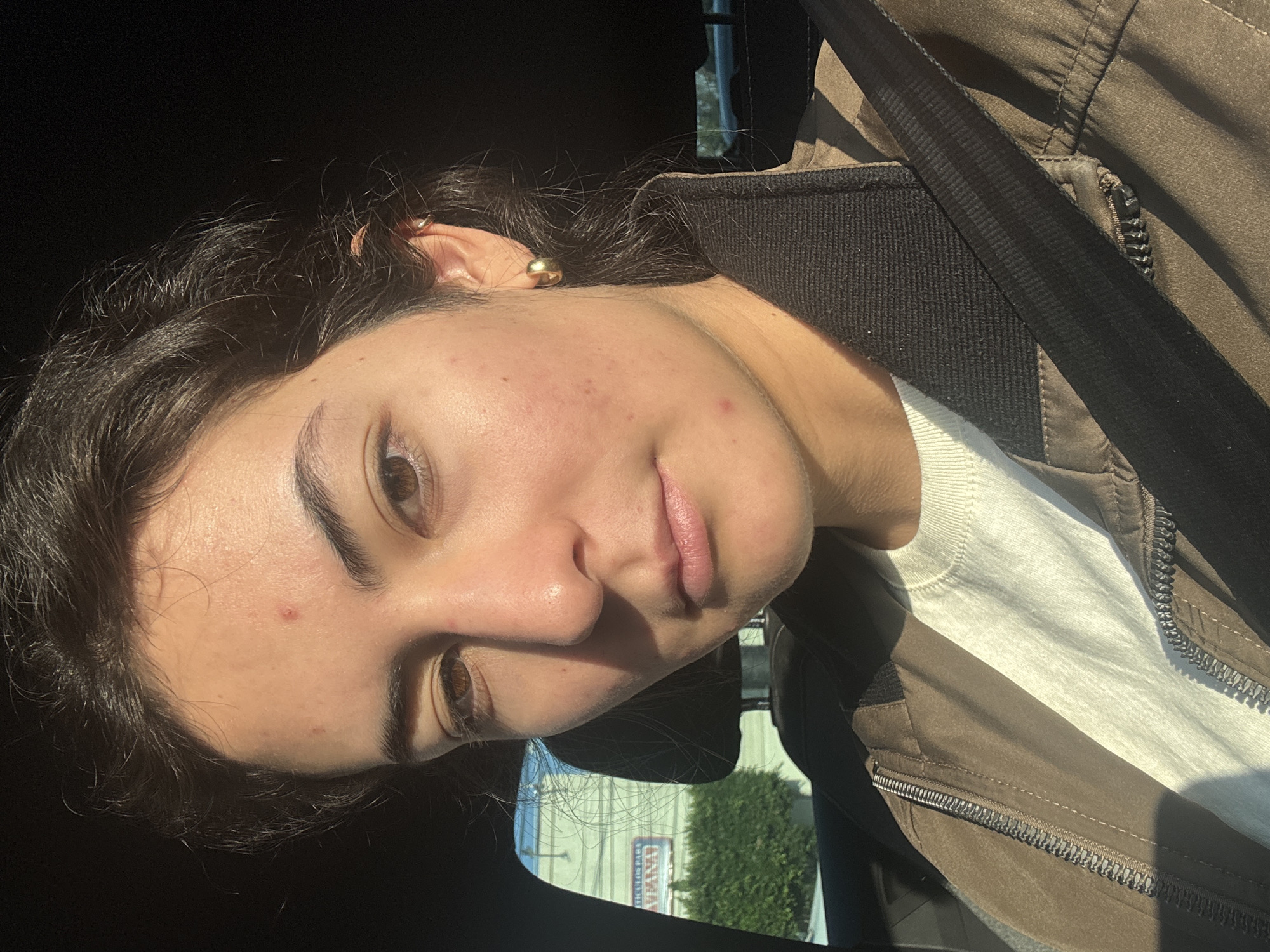 A medium shot of a young woman, likely in her late teens to early twenties, with brown eyes and dark, wavy hair pulled back. She is wearing a brown jacket over a white shirt and a seatbelt is visible across her body. She has a gold earring in her left ear. She has some acne spots on her face.
Through the car window, foliage and a building are visible. Based on the signage visible in the reflection, the location is Puebla, Mexico, possibly near a shop or business with the word "CANACO" on it.