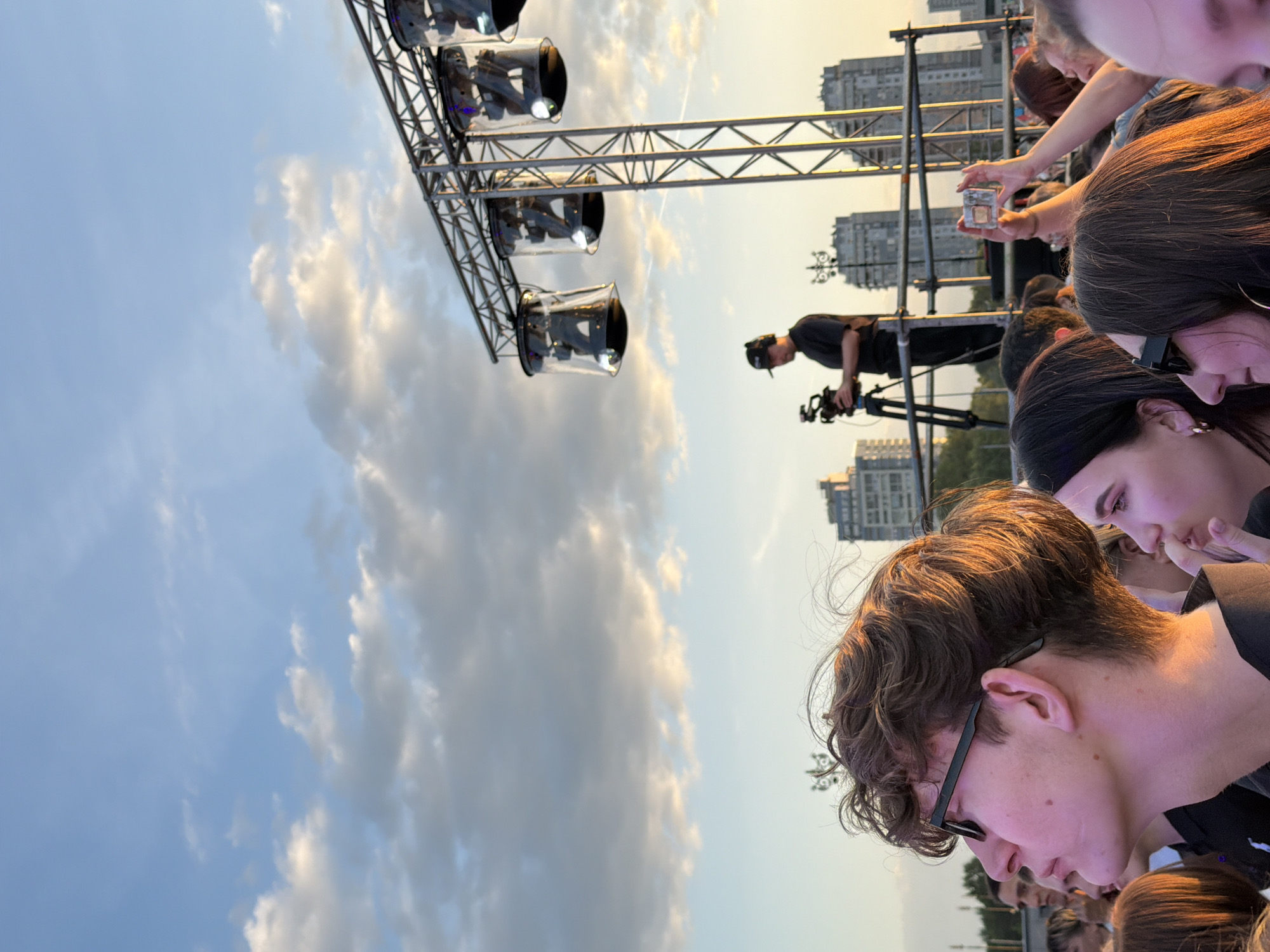 In an outdoor urban setting, a crowd of people are gathered, looking upwards towards a stage structure. The sky is blue with scattered clouds, and the sun is setting, casting a warm glow on the scene. A person in the foreground, wearing glasses, is looking up, with other faces partially visible around them. In the background, a person is operating a video camera on a tripod, positioned on a platform near the stage lights. Several large cylindrical lights hang from a metal truss structure. Buildings are visible in the distance, suggesting a city environment. One person in the crowd is holding up a phone, possibly to take a picture or record a video.
