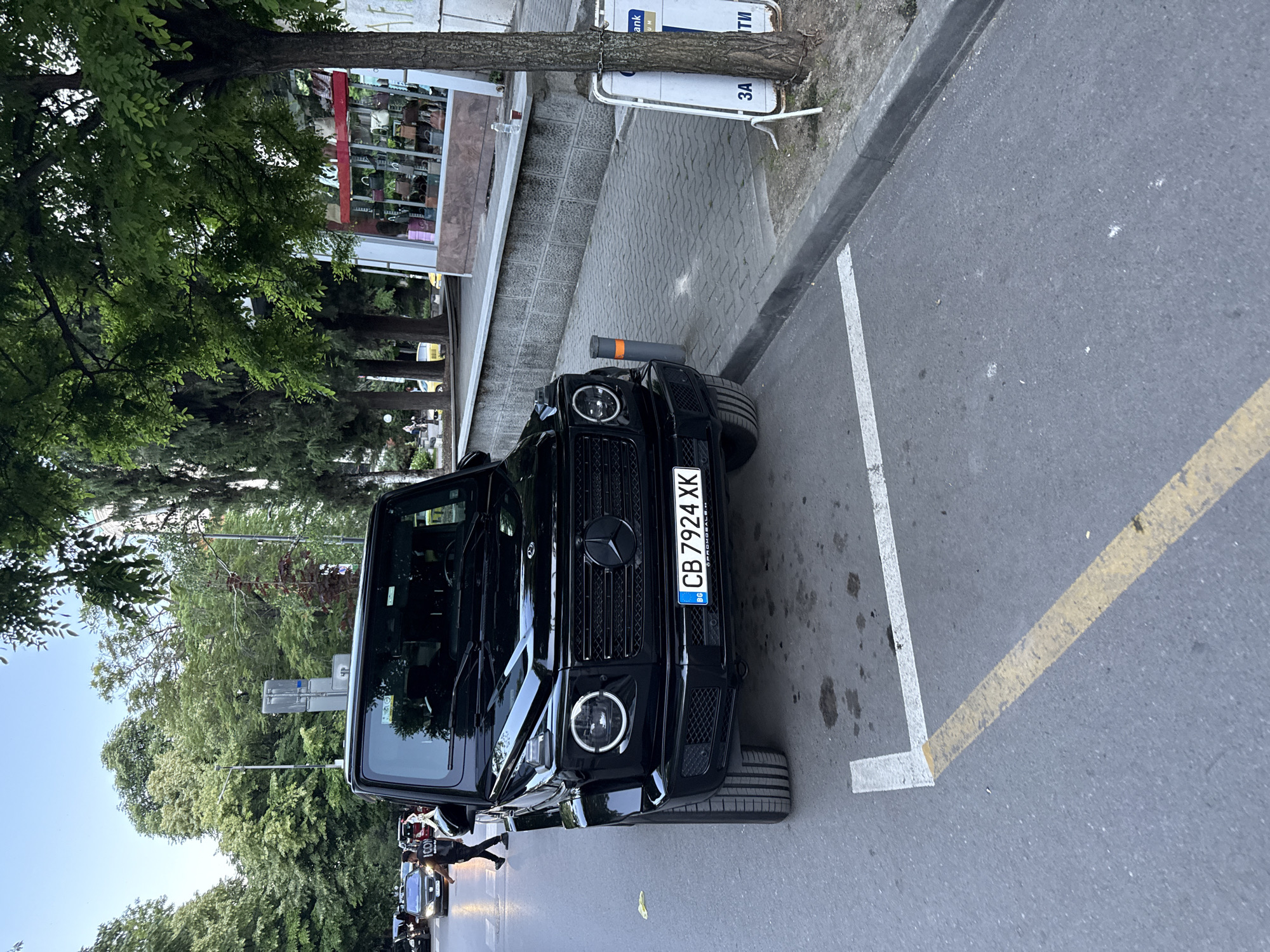 The image depicts a street scene in the city of Stara Zagora, Bulgaria. A black Mercedes-Benz G-Wagen is parked on the right side of the road, occupying a designated parking spot marked by white lines on the asphalt. The vehicle has a Bulgarian license plate reading "CB 7924 XK". The car is positioned in front of a building with a storefront, which displays various handbags.
The sidewalk adjacent to the road is paved with grey interlocking bricks. A tree with lush green foliage stands to the right of the parked car, partially obscuring the building. A light pole is visible further down the street. Across the street, several other vehicles are present, and pedestrians are walking. The sky is clear and appears to be late afternoon or early evening. The overall environment suggests a commercial or residential area within the city.