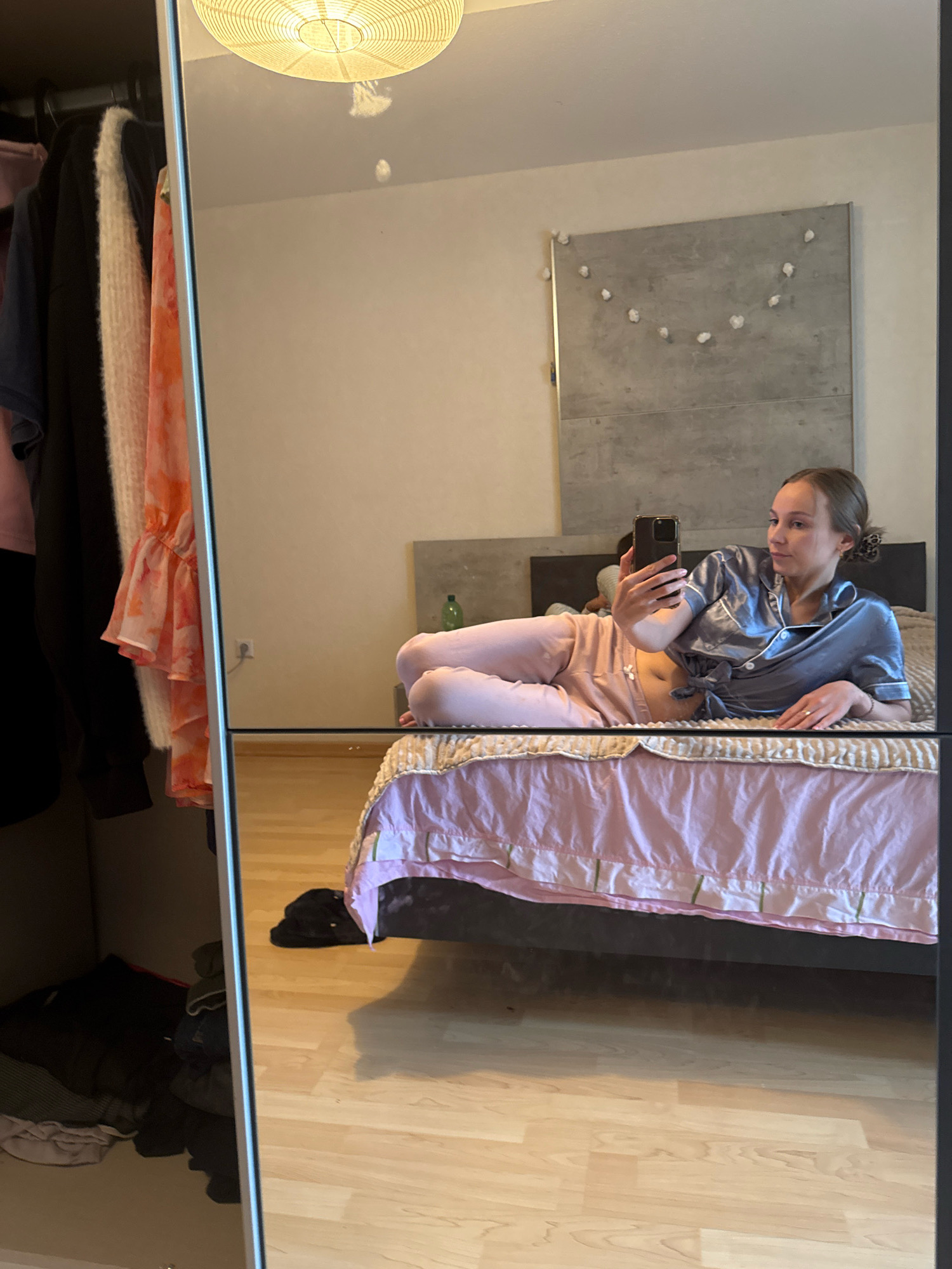 In a bedroom in Überherrn, Germany, a young woman is lying on a bed, holding a smartphone and taking a mirror selfie. She is wearing blue satin pajamas and pink lounge pants. The bed is covered with a pink duvet and a white comforter. To the left of the bed, a closet is visible, filled with clothing. The room has light-colored wooden flooring and a grey headboard on the bed. A wall with a concrete panel and a string of decorative lights hangs in the background. A light fixture is visible on the ceiling.