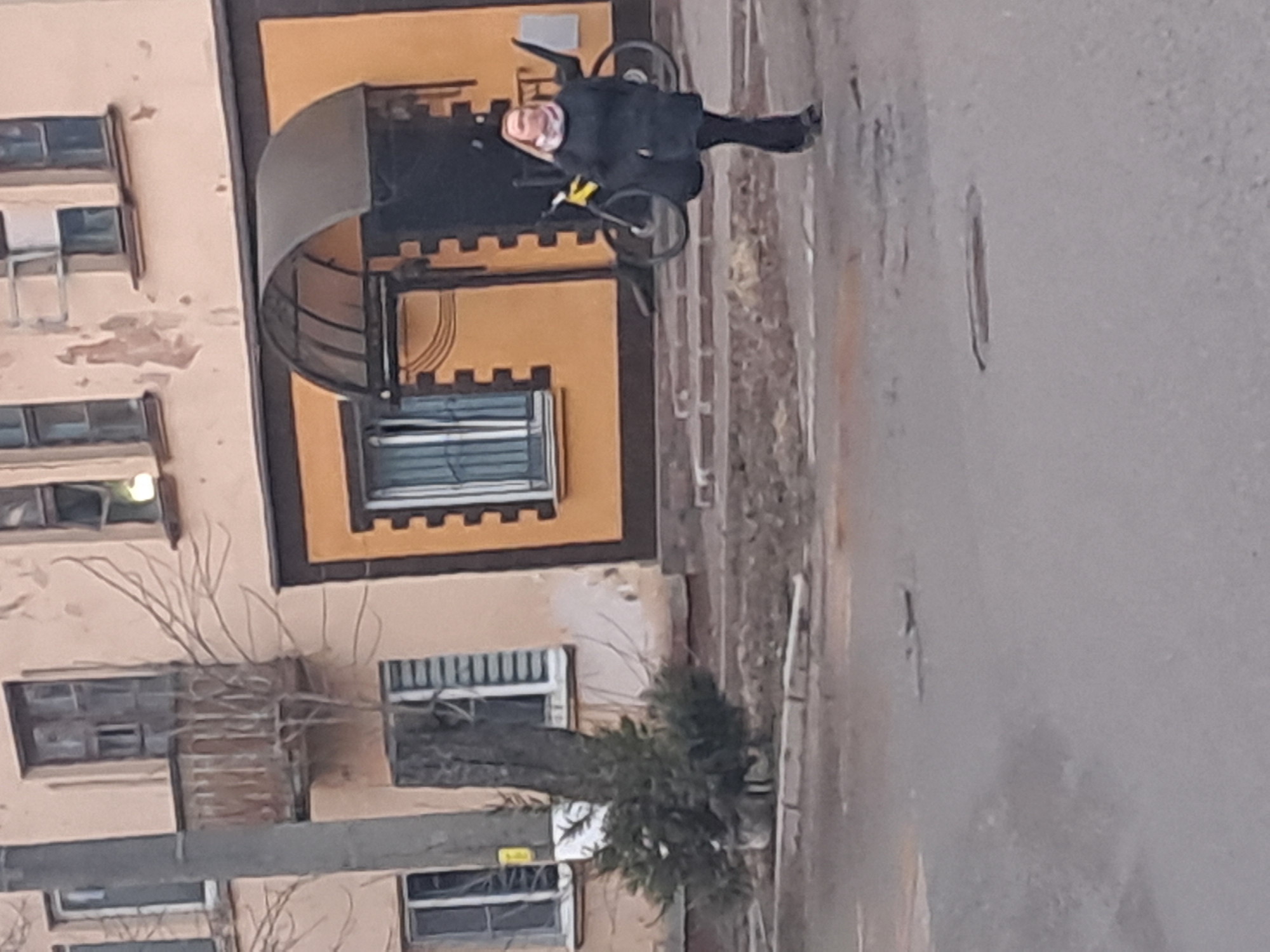The image portrays an outdoor urban scene in Elektrostal, Russia, observed under diffuse daylight conditions. The foreground consists of a grey asphalt surface, exhibiting multiple minor cracks and patches. A single individual with light-colored hair is captured in motion, walking leftward across the mid-ground. This person is attired in a dark long coat, dark trousers, and a light-colored neck scarf, and is carrying a dark bag over their left shoulder.
Behind the individual, a concrete curb separates the asphalt from a dirt and gravel strip. Positioned within this strip, slightly left of center, is a tall, bare tree trunk, at the base of which a small evergreen bush grows. To the left of the scene, a multi-story building, predominantly light peach in color, displays areas of peeling paint on its facade. It features multiple rectangular windows with light frames and a weathered wooden balcony with a railing on an upper level.
Adjacent to this building on the right, another structure features a contrasting lower section painted orange with dark, segmented trim outlining its windows and main entrance. A dark, arched canopy extends over this entrance. A yellow bicycle with dark tires is leaning against the orange facade, positioned to the right of the entrance. The upper portion of this second building matches the light peach color and weathered appearance of the adjacent structure.