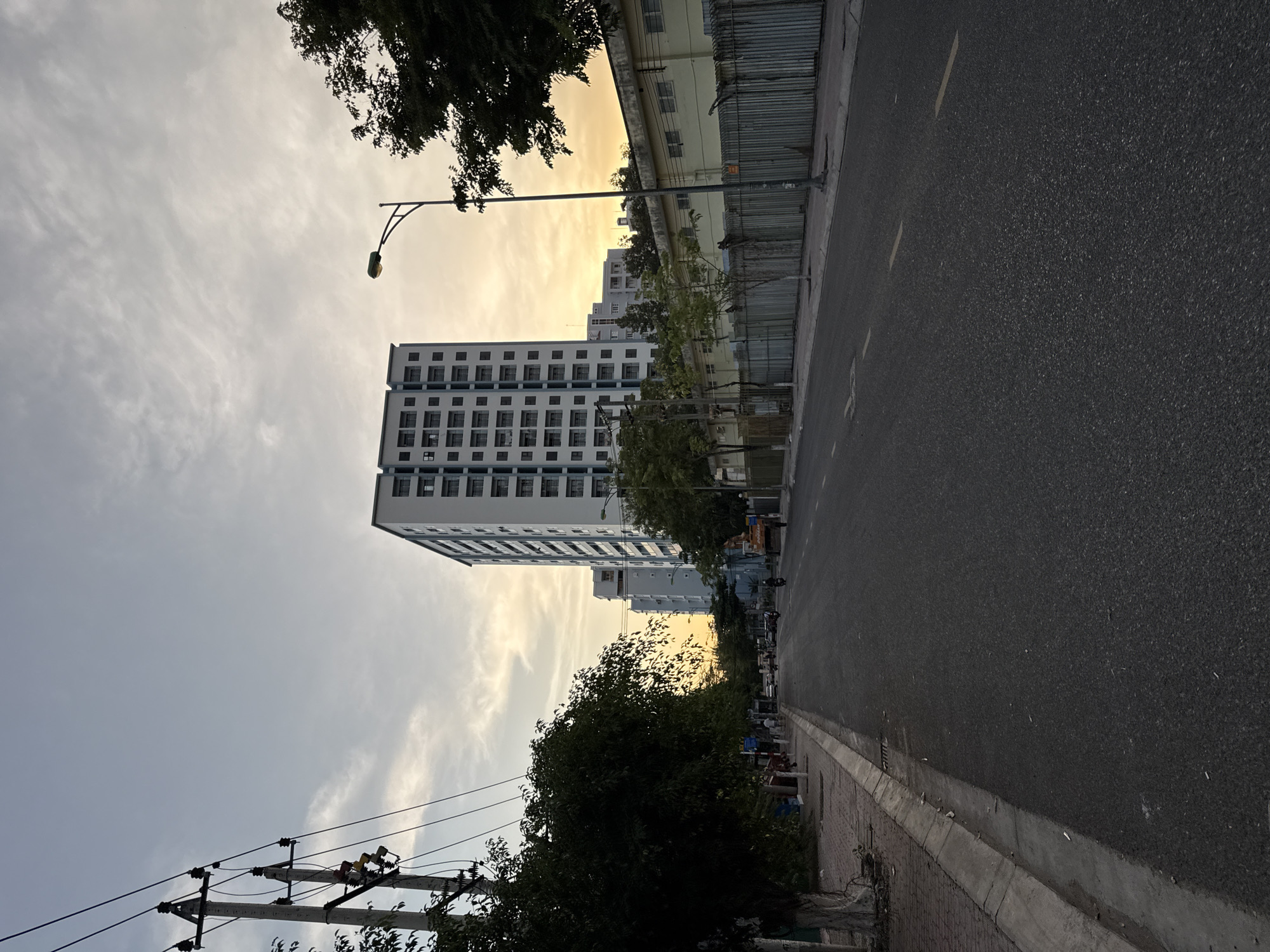 The image depicts a street scene in Nha Trang, Vietnam, at what appears to be dusk. A paved road runs through the center, flanked by a sidewalk on the left and a metal fence with a building behind it on the right. Tall residential buildings with multiple windows are visible in the background. A street lamp stands on the right side of the road. Power lines and a utility pole are prominent on the left. A few vehicles and pedestrians are present on the street and sidewalk, suggesting moderate activity. The sky is overcast with hints of sunset colors.