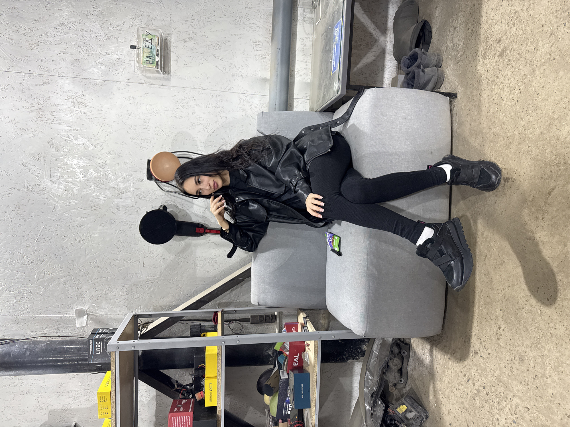 A young woman with long, dark hair is lying casually on a light grey sofa, looking directly at the camera with a subtle smile. She is dressed in a black leather jacket, black leggings, white socks, and black chunky sneakers.
The setting appears to be an informal indoor space, possibly a workshop, studio, or a casual storage area. The wall behind her features a light-colored, textured surface, resembling oriented strand board. Affixed to the wall is a clear plastic box containing paper currency, with the text "НА ЕДУ" (meaning "FOR FOOD" in Russian) and "200" visible. Near the woman's head, a dark, round object, possibly a microphone or part of a stand, is present. A small green and purple item is tucked into the sofa cushion near her hip.
To the right of the sofa, on the concrete floor, a pair of grey, fur-lined slippers or boots are placed. To the left, a metal shelving unit is filled with various items, including yellow boxes labeled "LITEC" and "LED," red boxes marked "EAL," and a blue box displaying "SEE THE FUTURE." A dark pipe or conduit runs horizontally in the upper background. The floor is a rough concrete surface. The location context is Chaikovsky, Russia.